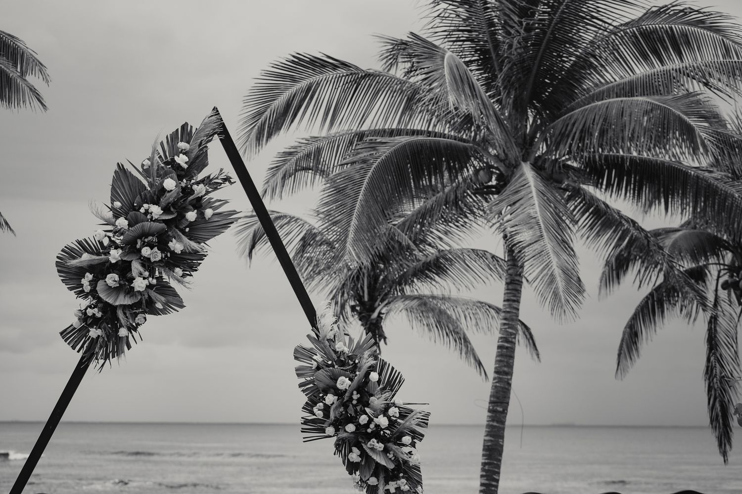 Black and white beach scene with palm trees and decorated poles by the ocean shore.
