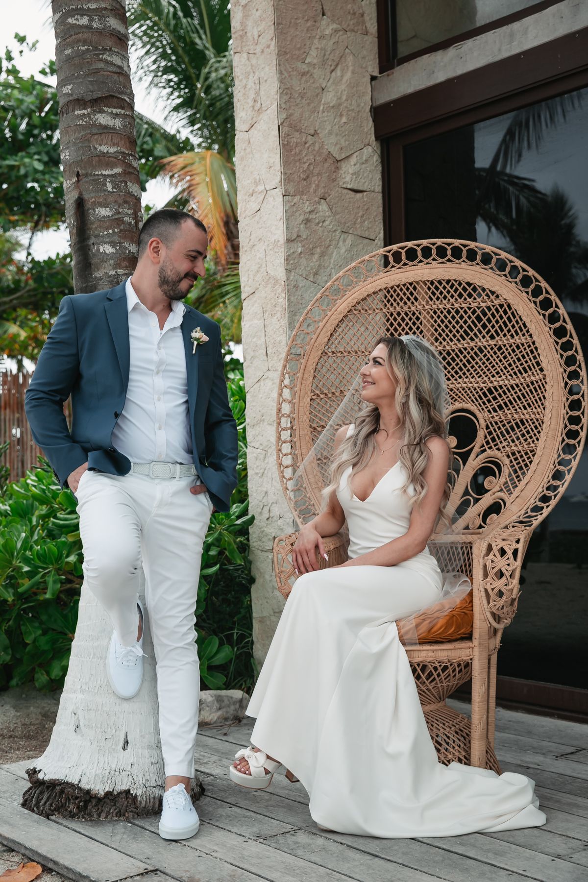 Bride in white gown seated in rattan peacock chair smiling at groom in blue blazer outdoors.