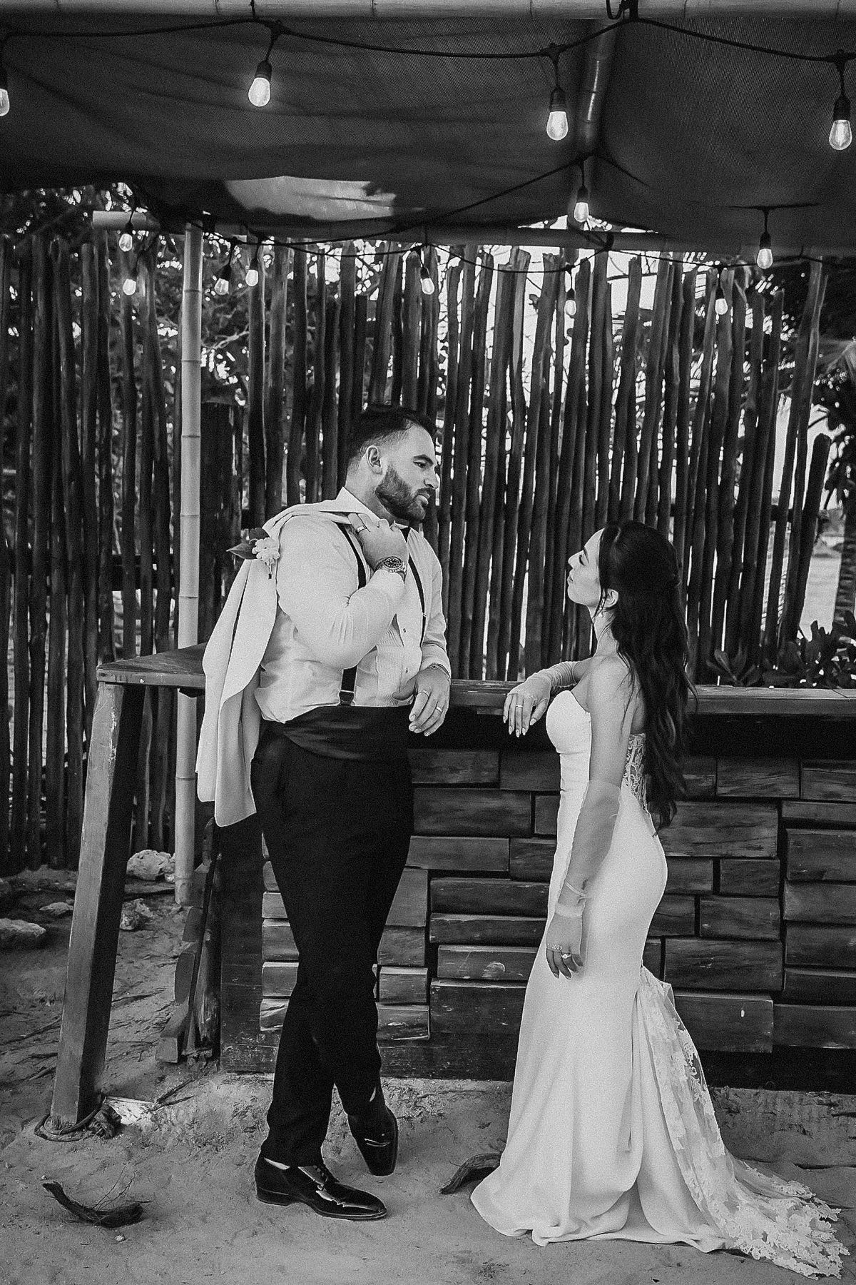 Black and white photo of a bride in a fitted gown and groom in suspenders sharing a romantic moment at a rustic outdoor venue.