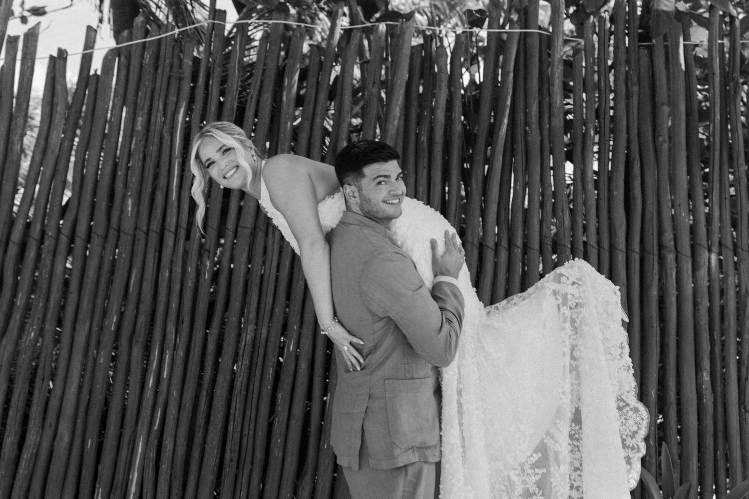 Black and white photo of a groom carrying his bride in a wedding dress near a rustic wooden fence backdrop.