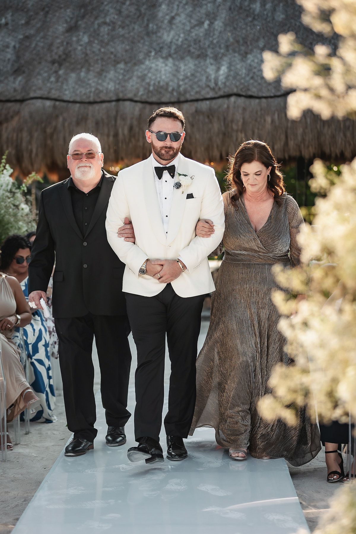 Groom in white tuxedo jacket walks down wedding aisle escorted by parents at outdoor tropical ceremony.