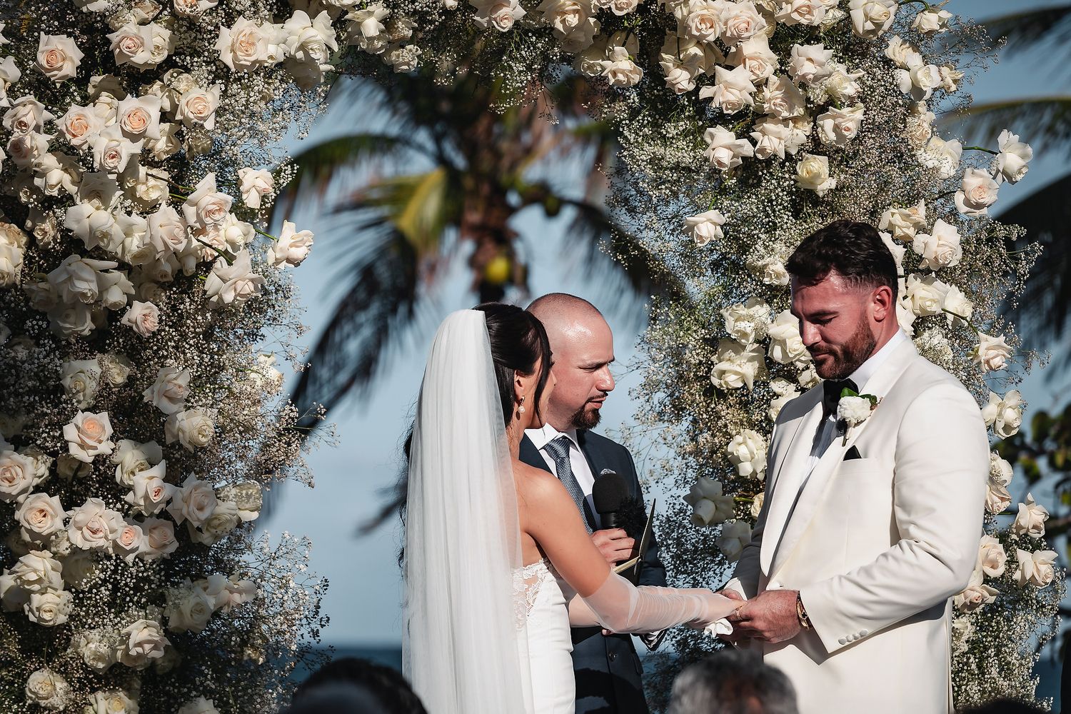 Bride and groom exchange vows under a floral arch with white roses at an outdoor wedding ceremony.