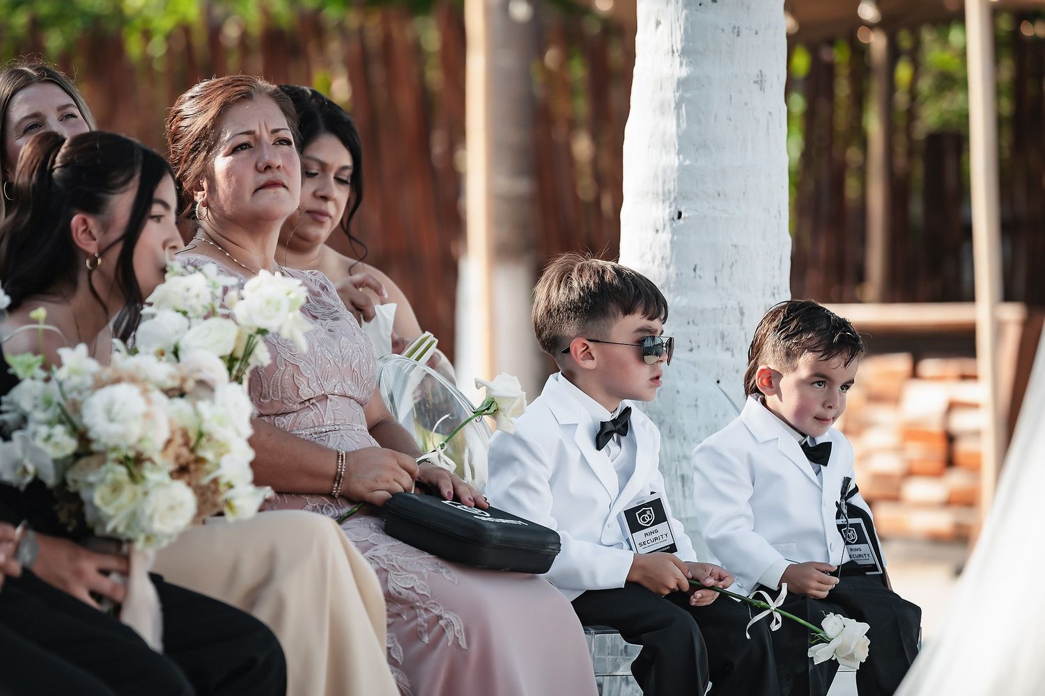 Wedding guests seated at outdoor ceremony, including woman in pink gown and two boys in white tuxedos with sunglasses.