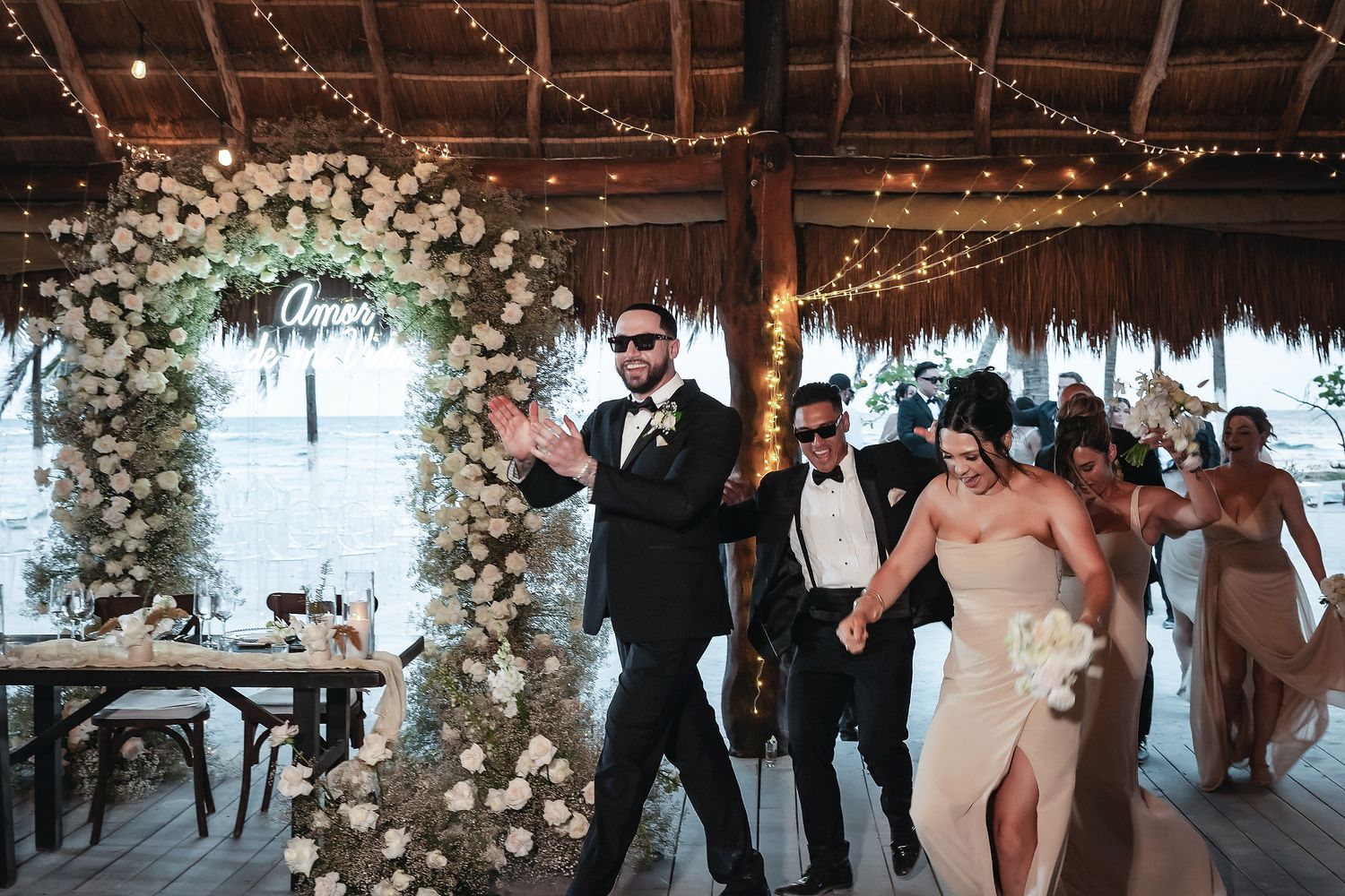 Wedding couple makes a joyful grand entrance under a floral arch with white roses at a tropical beachside venue.