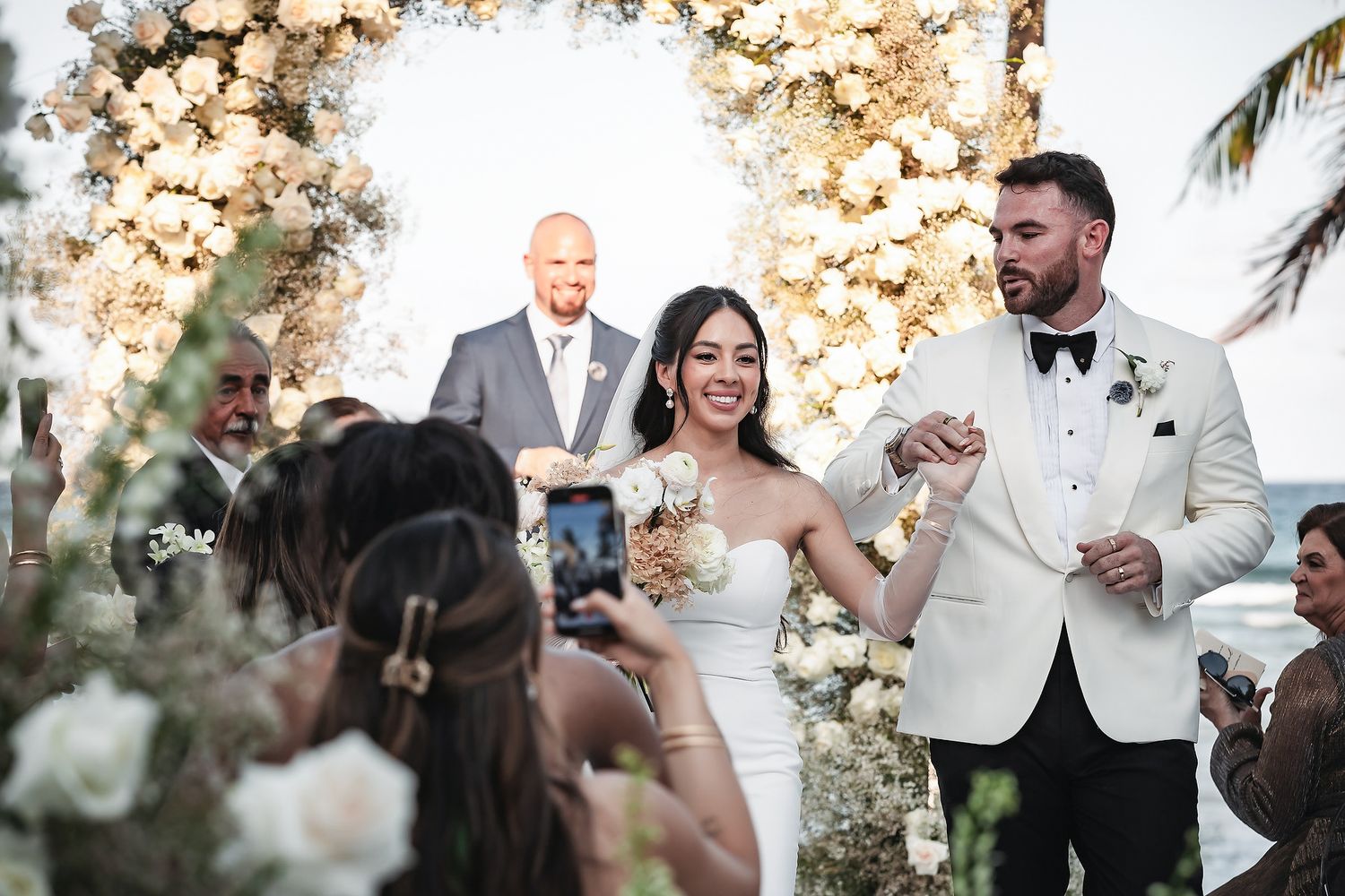 Bride and groom smiling and holding hands at outdoor wedding ceremony with floral arch and guests.