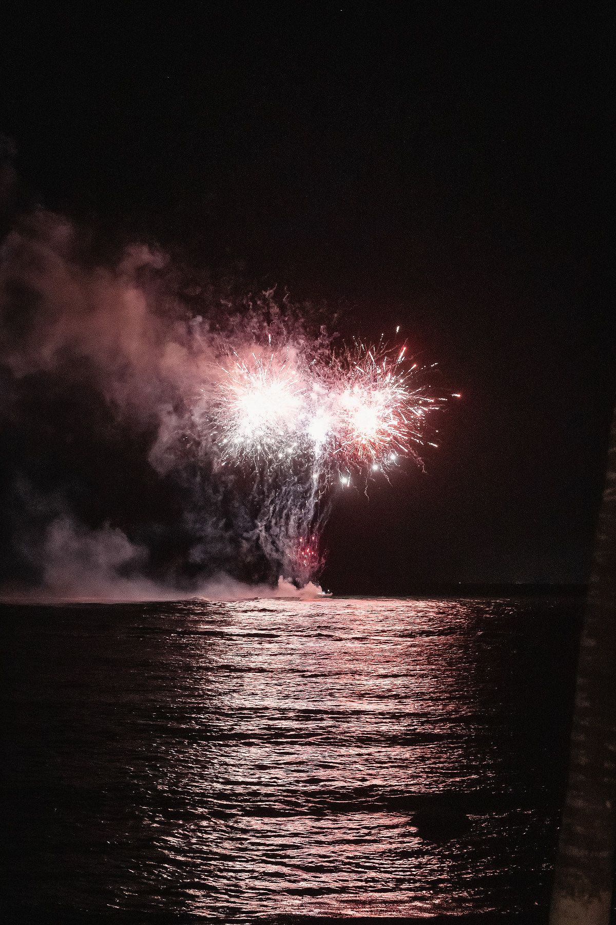 Colorful fireworks exploding over water at night, reflecting pink and red light on the ocean surface.