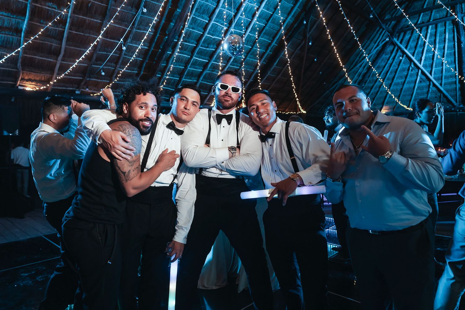 Group of groomsmen pose together smiling at a wedding reception inside a rustic barn venue with string lights.