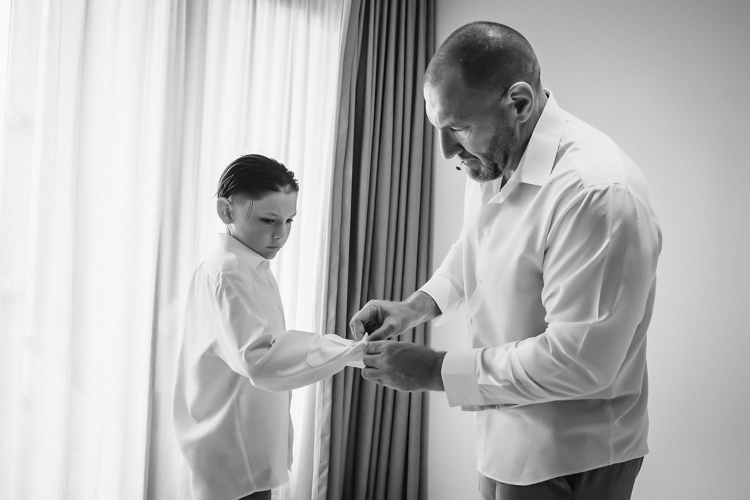 Man helping young boy button his shirt cuffs in black and white wedding preparation photo.