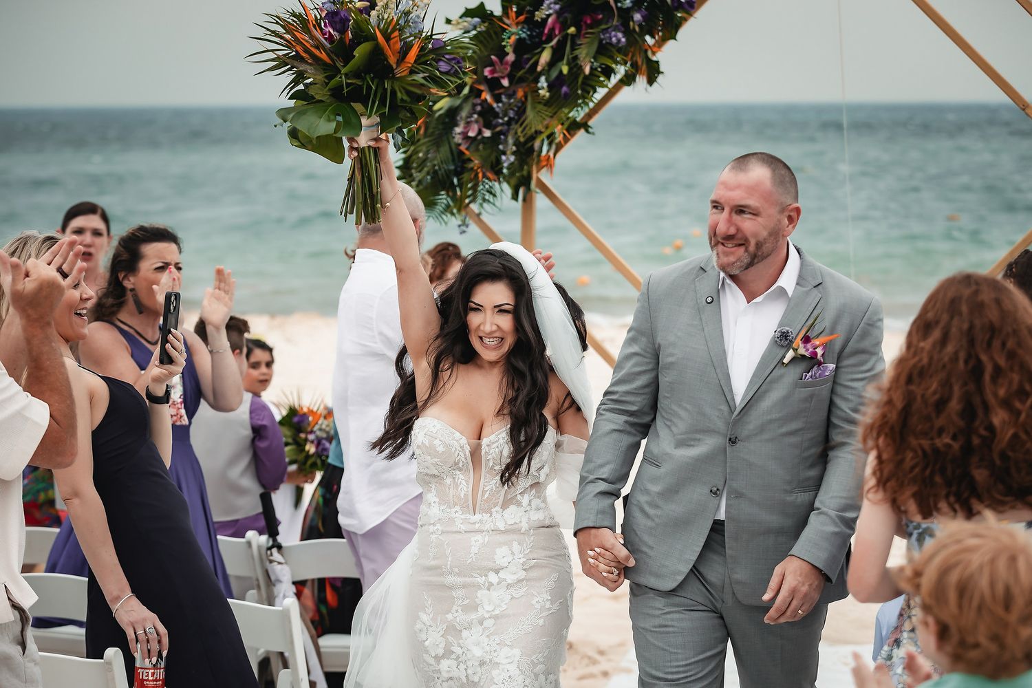 Bride and groom walk down beach aisle after wedding ceremony, bride raising bouquet in celebration.