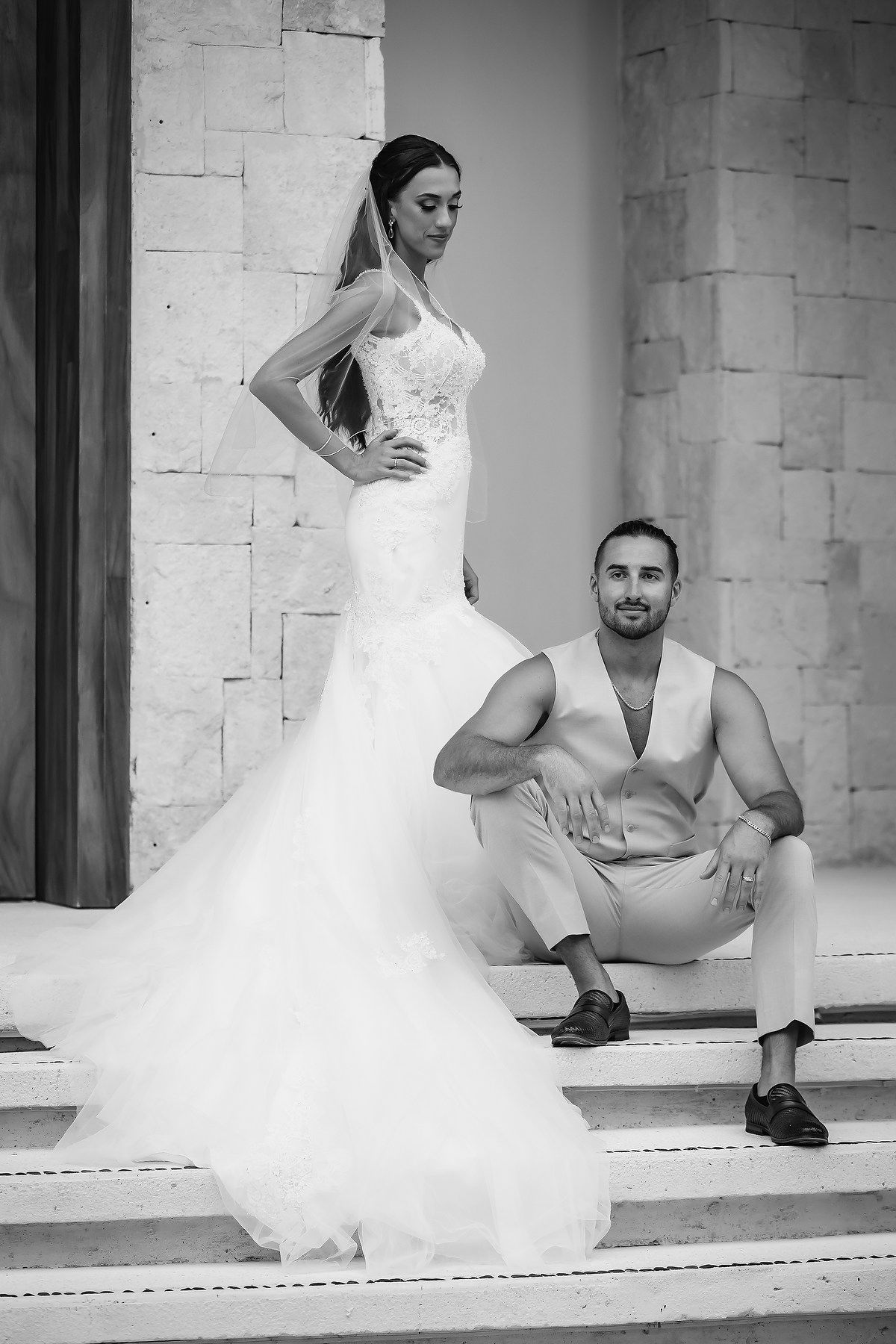 Black and white photo of a bride in a mermaid gown standing beside a seated groom on elegant stone steps.