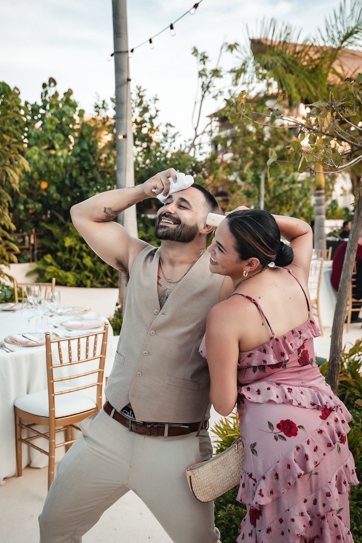 A smiling couple in elegant attire posing playfully at an outdoor wedding reception surrounded by lush greenery.