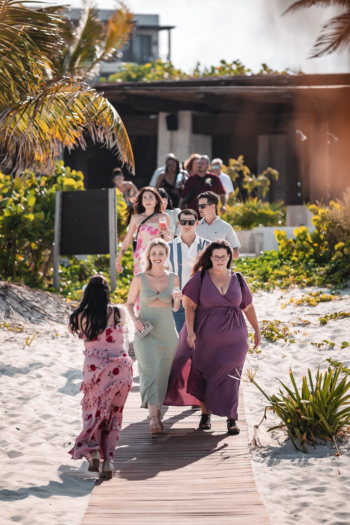 Group of guests in colorful maxi dresses walking along a wooden boardwalk path at a tropical beach resort.