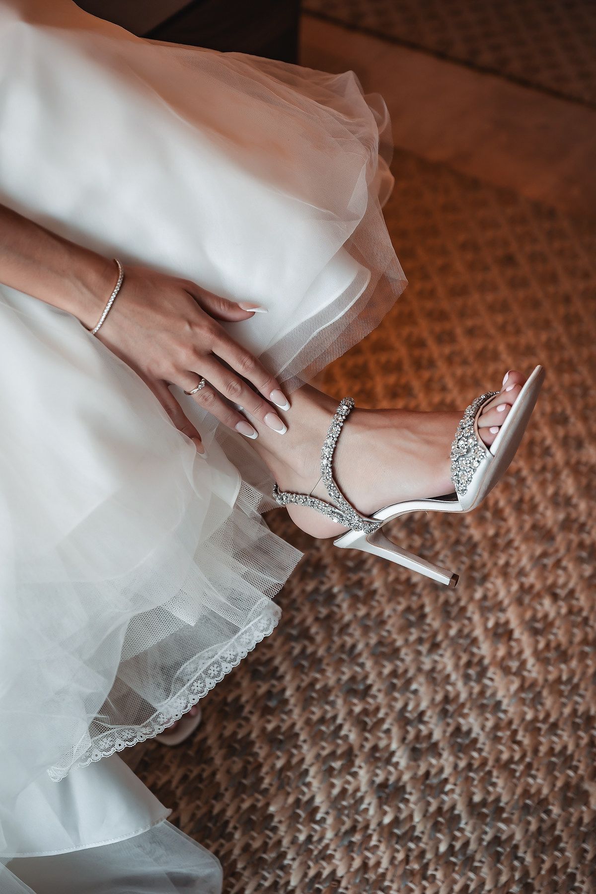 Bride wearing sparkling crystal embellished white stiletto heels with ankle strap, adjusting shoe under flowing wedding dress.