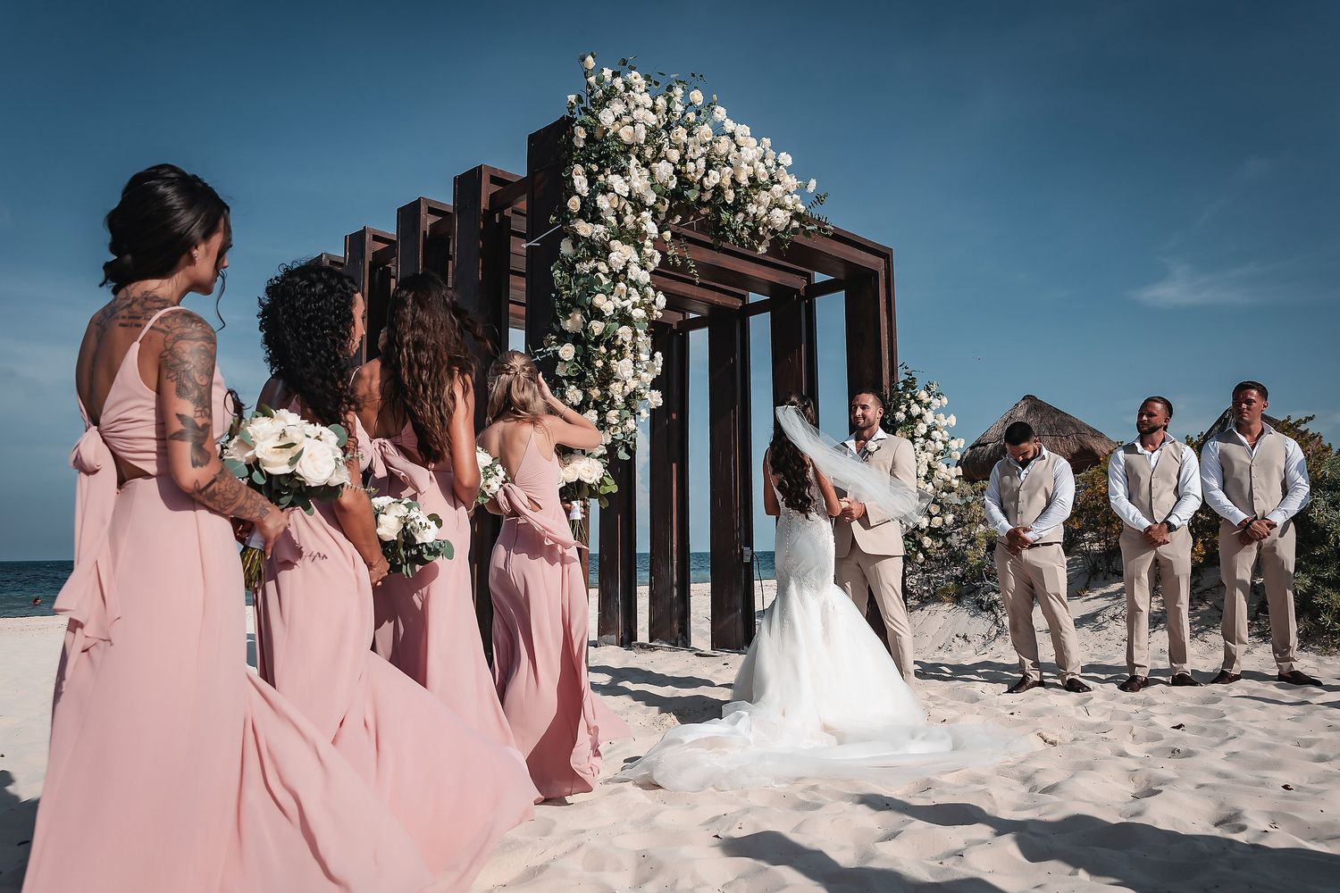 Beach wedding ceremony with bridesmaids in pink dresses and groomsmen beside a floral arch on white sand.