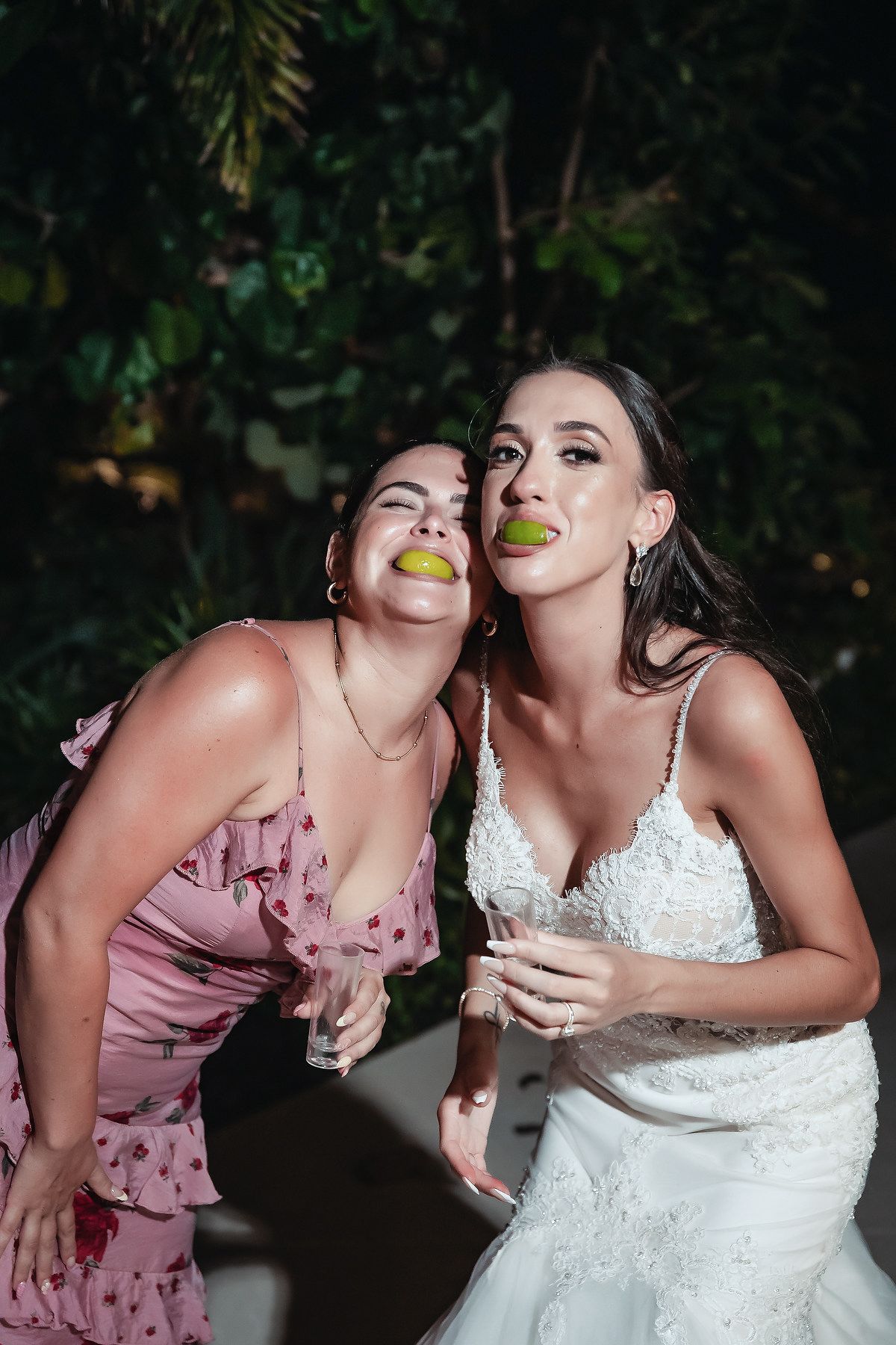 Bride and friend playfully pose with limes in their mouths during fun outdoor wedding reception celebration.