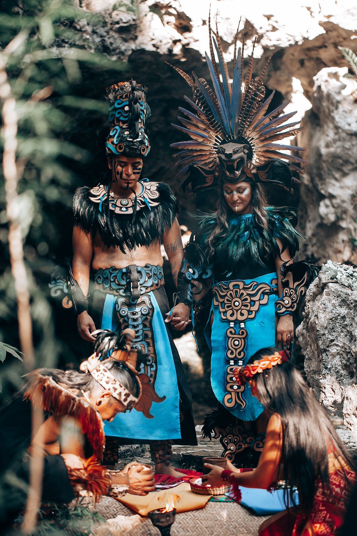 Two Mayan-costumed performers in blue feathered headdresses during traditional ground ceremony outdoors.