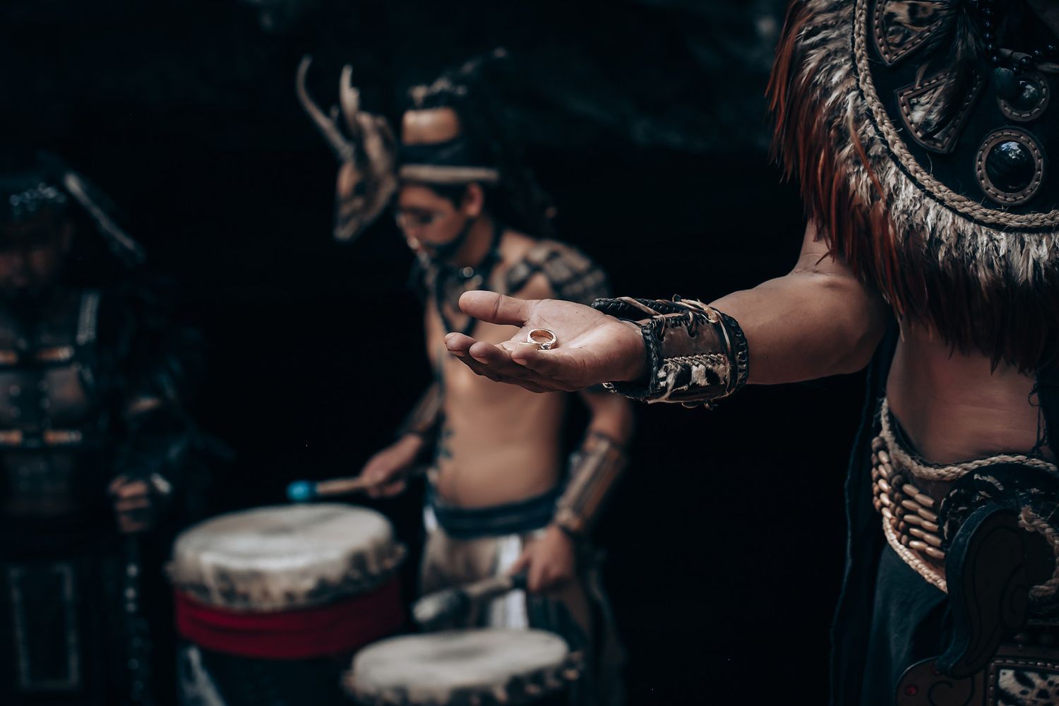Close-up of performer in Mayan costume extending hand while drummer plays in dark background.