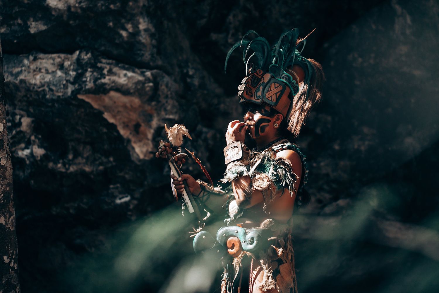 Mayan warrior in traditional ceremonial costume with feathered headdress holding artifact in rocky jungle setting.