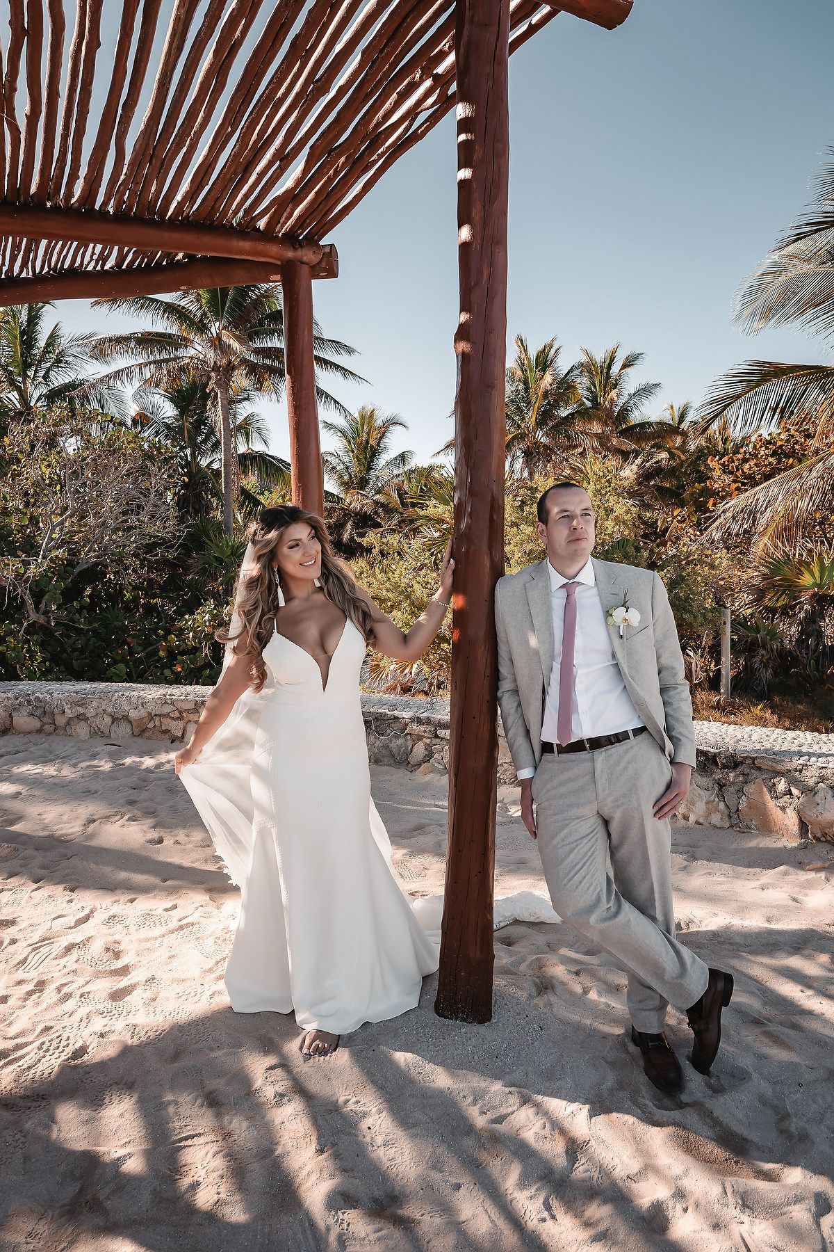 Bride and groom pose on sandy beach under rustic wooden pergola with tropical palm trees in background.