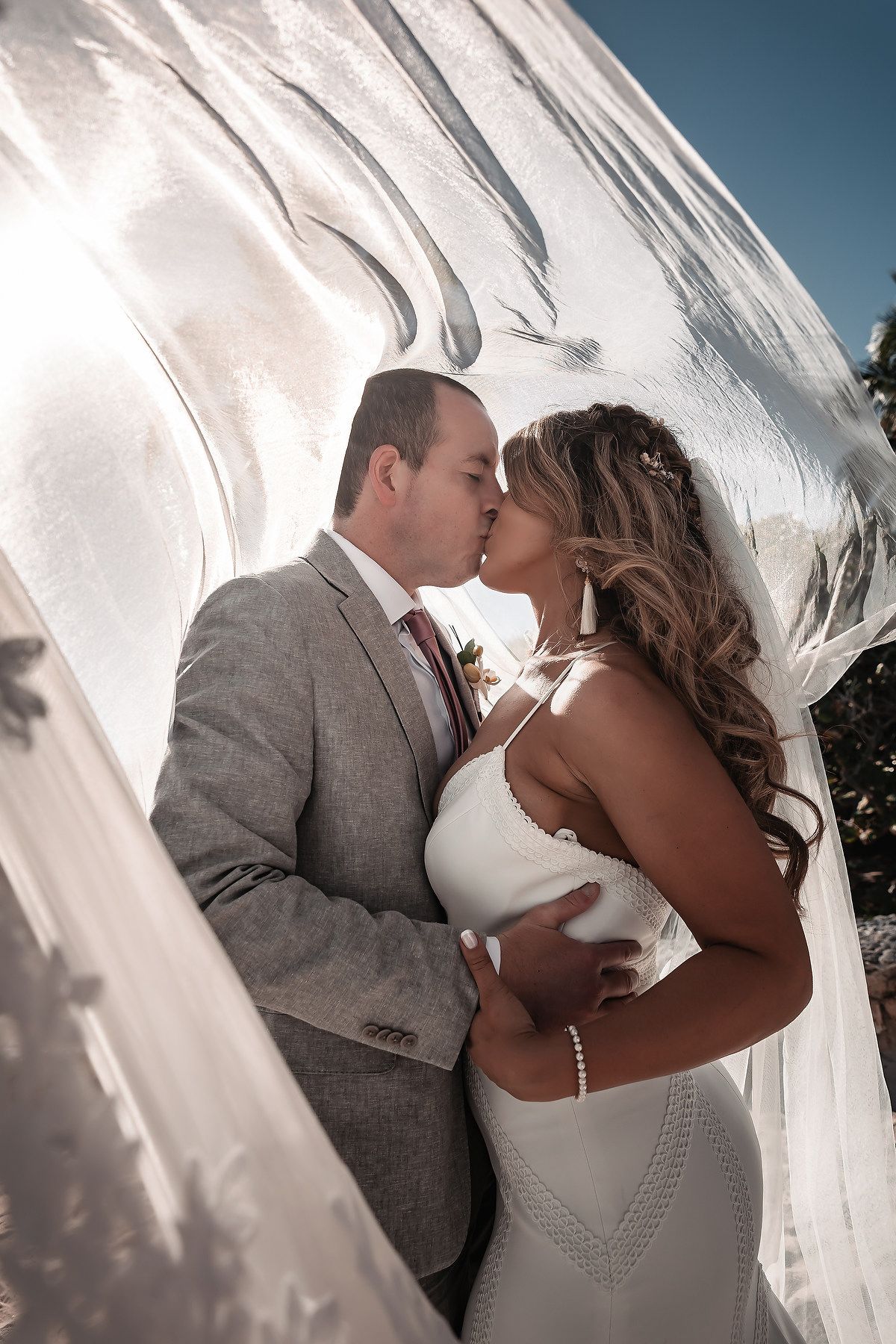 Bride and groom sharing a romantic kiss under a flowing white veil, backlit by sunlight on their wedding day.