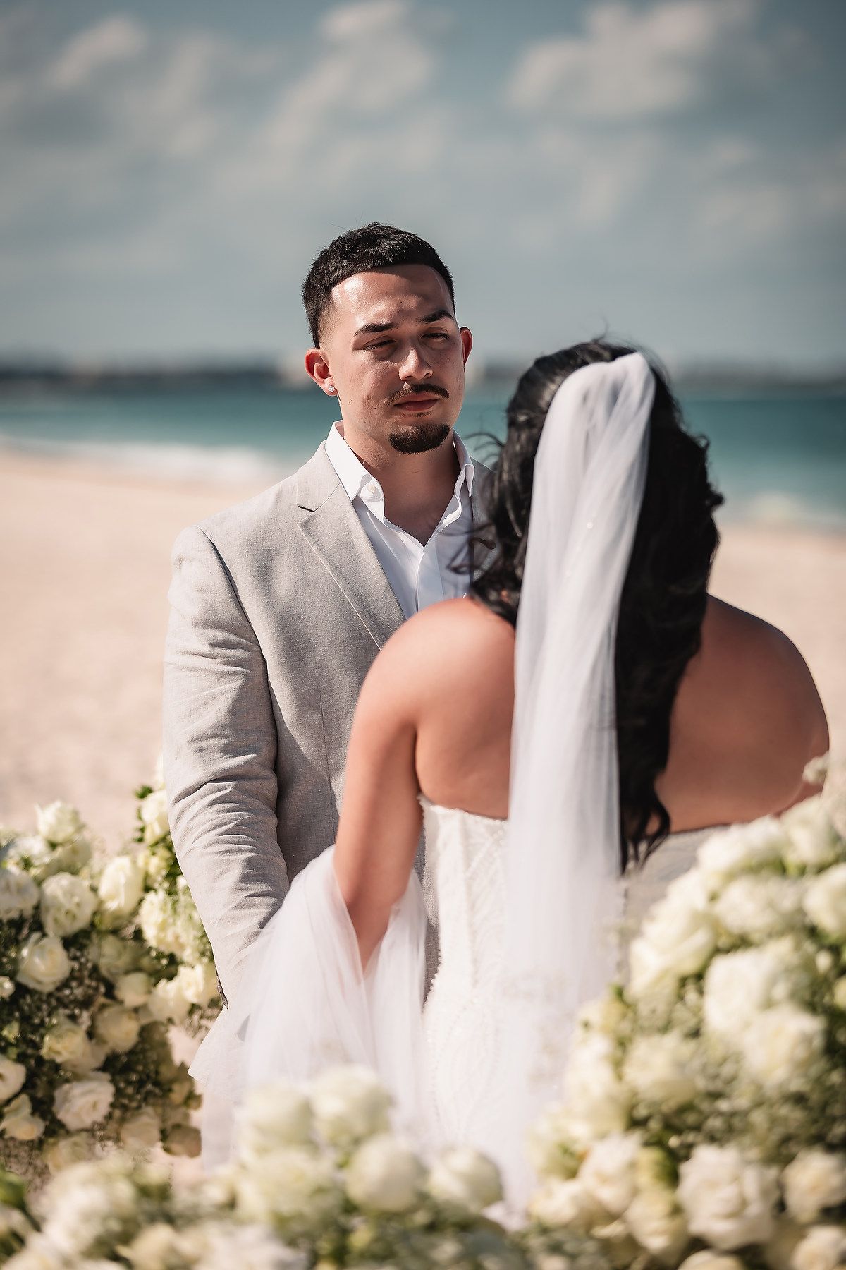 Groom in gray suit gazes at bride in white gown during beach wedding ceremony with white floral decor.