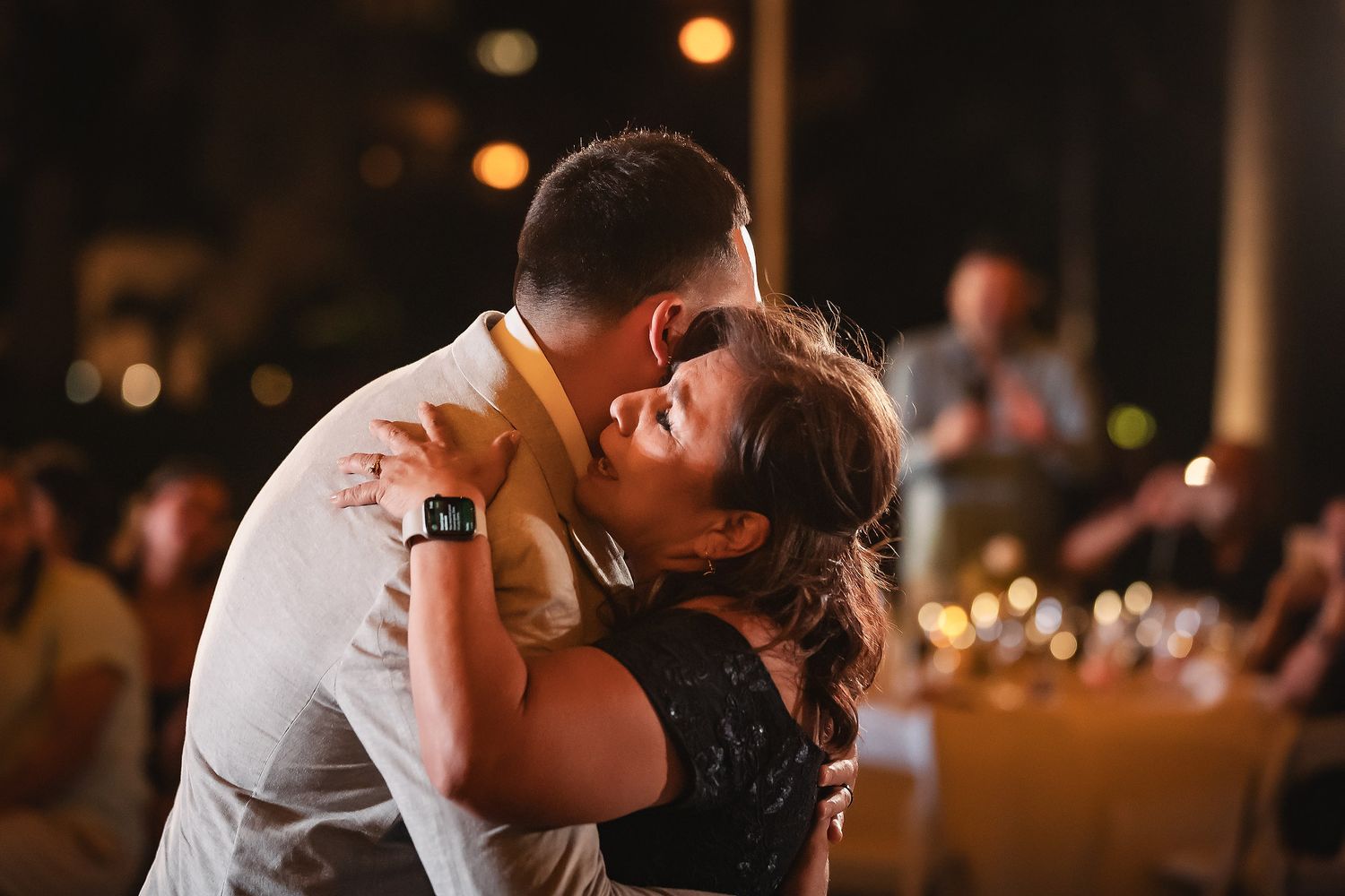 A couple shares a romantic dance embrace at a wedding reception with guests and warm lighting in the background.