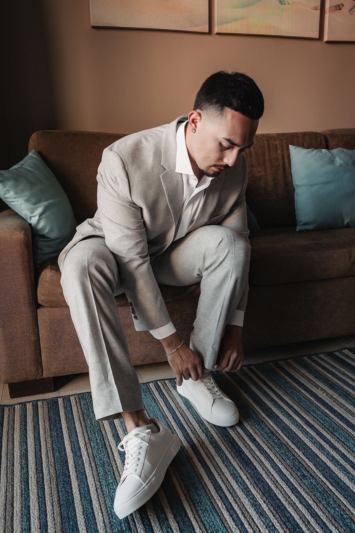 Man in light gray suit tying white sneakers while sitting on brown sofa with teal pillows.