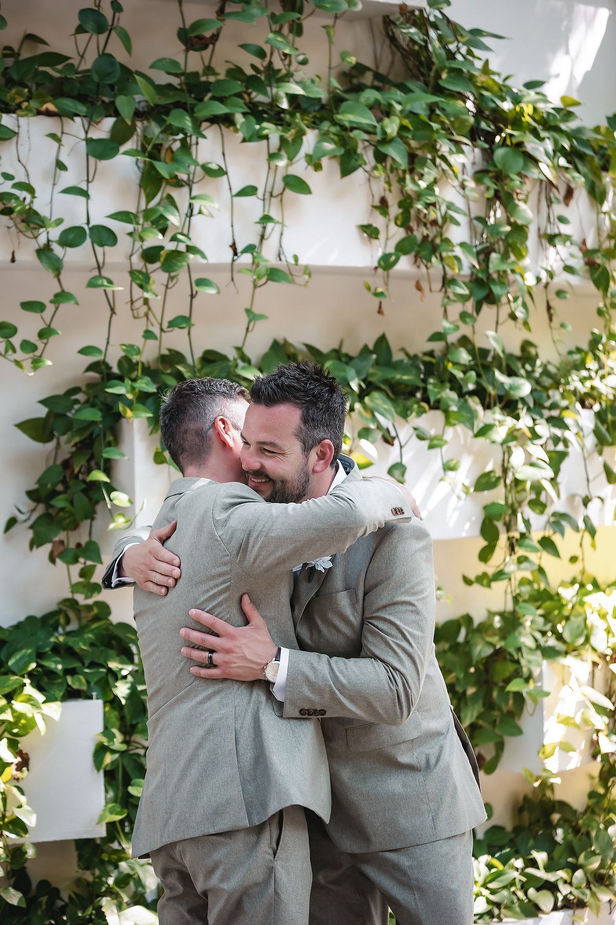 Two grooms in gray suits embrace and smile during their wedding ceremony surrounded by lush green ivy.