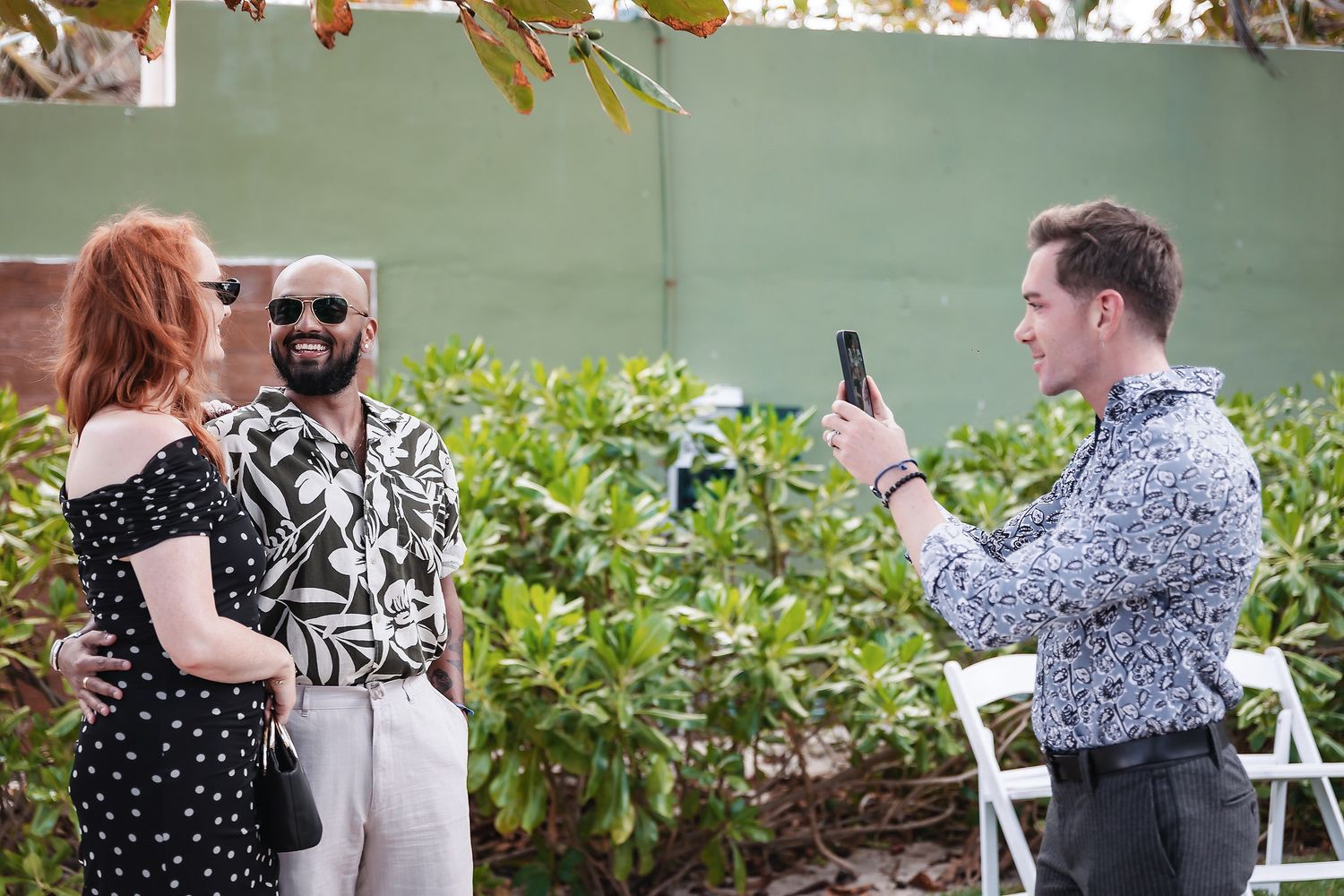 Man photographing smiling couple with smartphone at an outdoor tropical wedding ceremony venue.