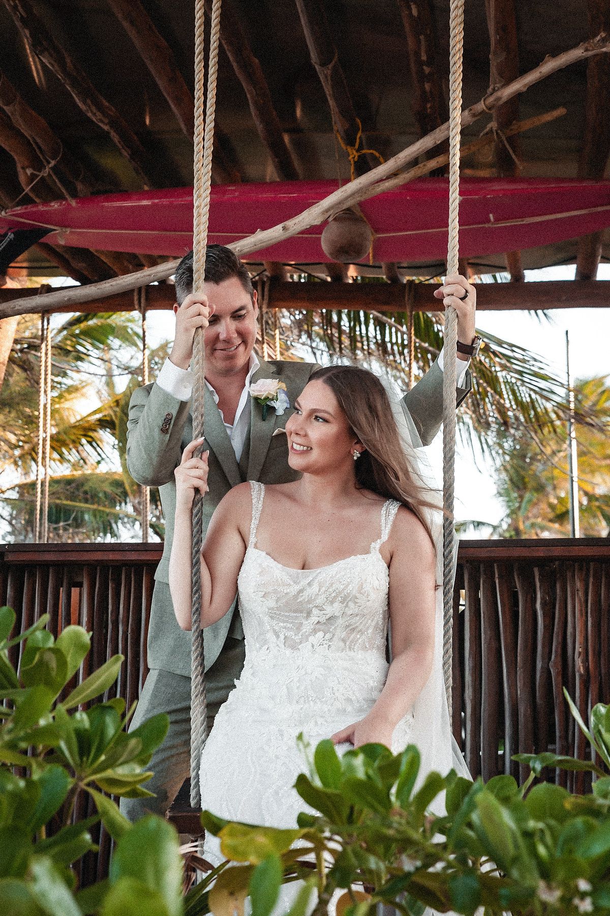 Bride in white gown sits on rope swing while partner in gray suit stands behind her at tropical wedding venue.