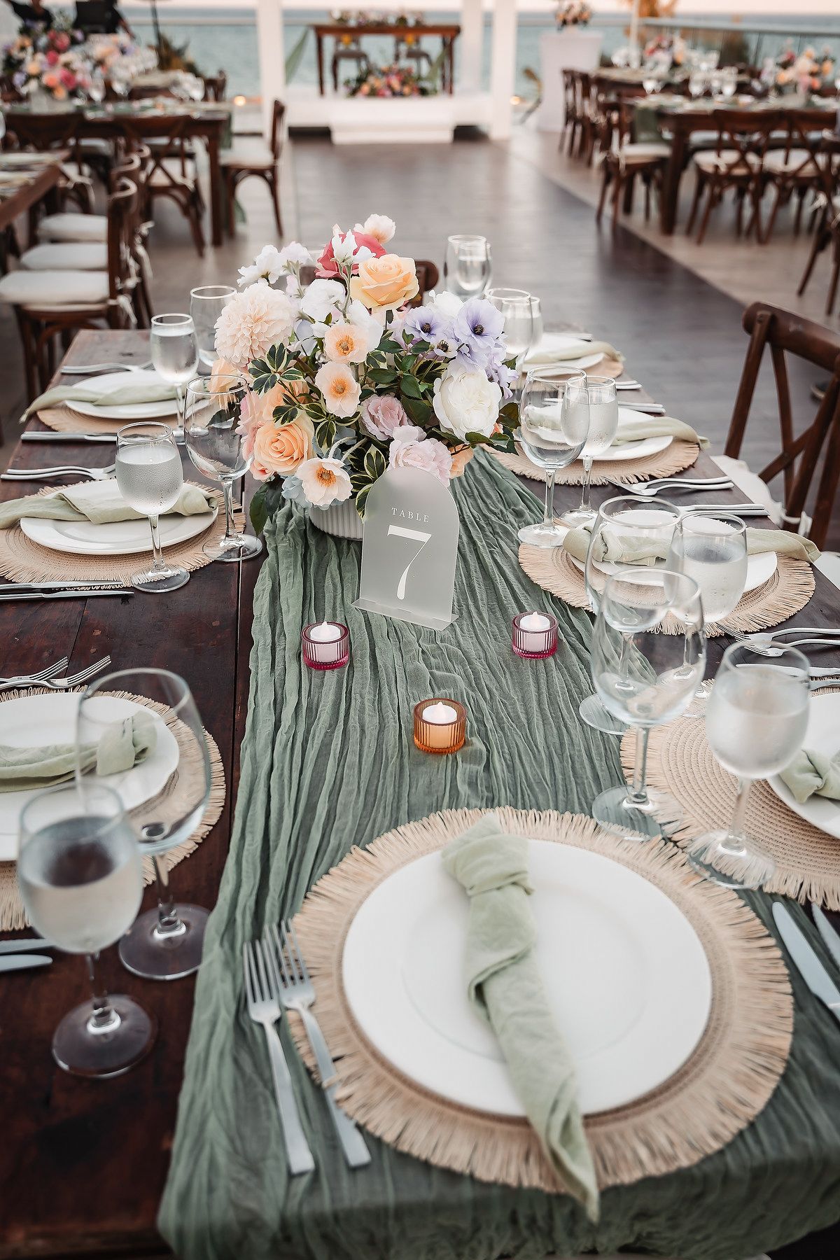 Elegant wedding reception table with sage green runner, woven chargers, floral centerpiece, and candles.
