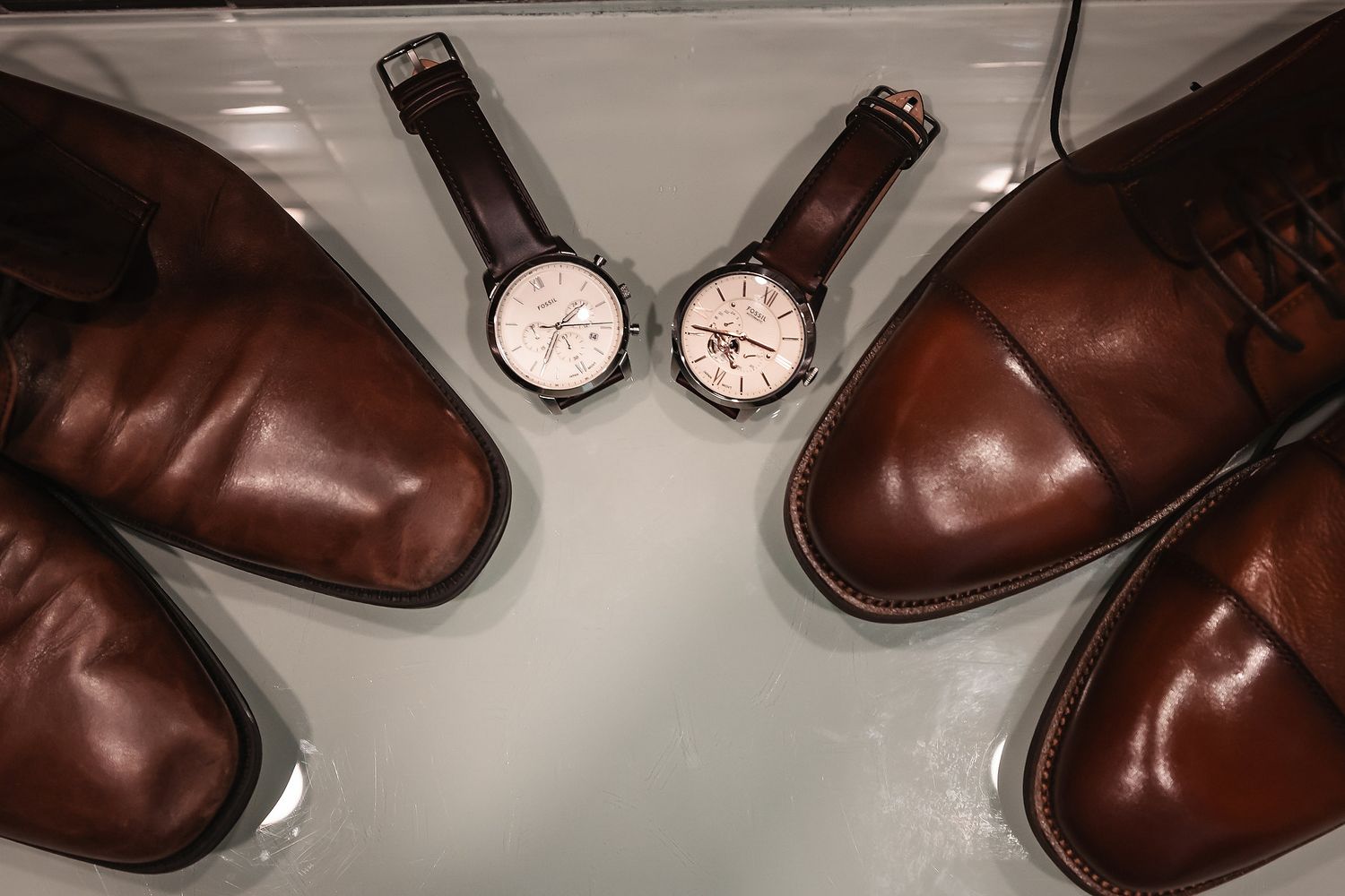 Brown leather dress shoes flanking two analog wristwatches on a white surface, groom wedding accessories.