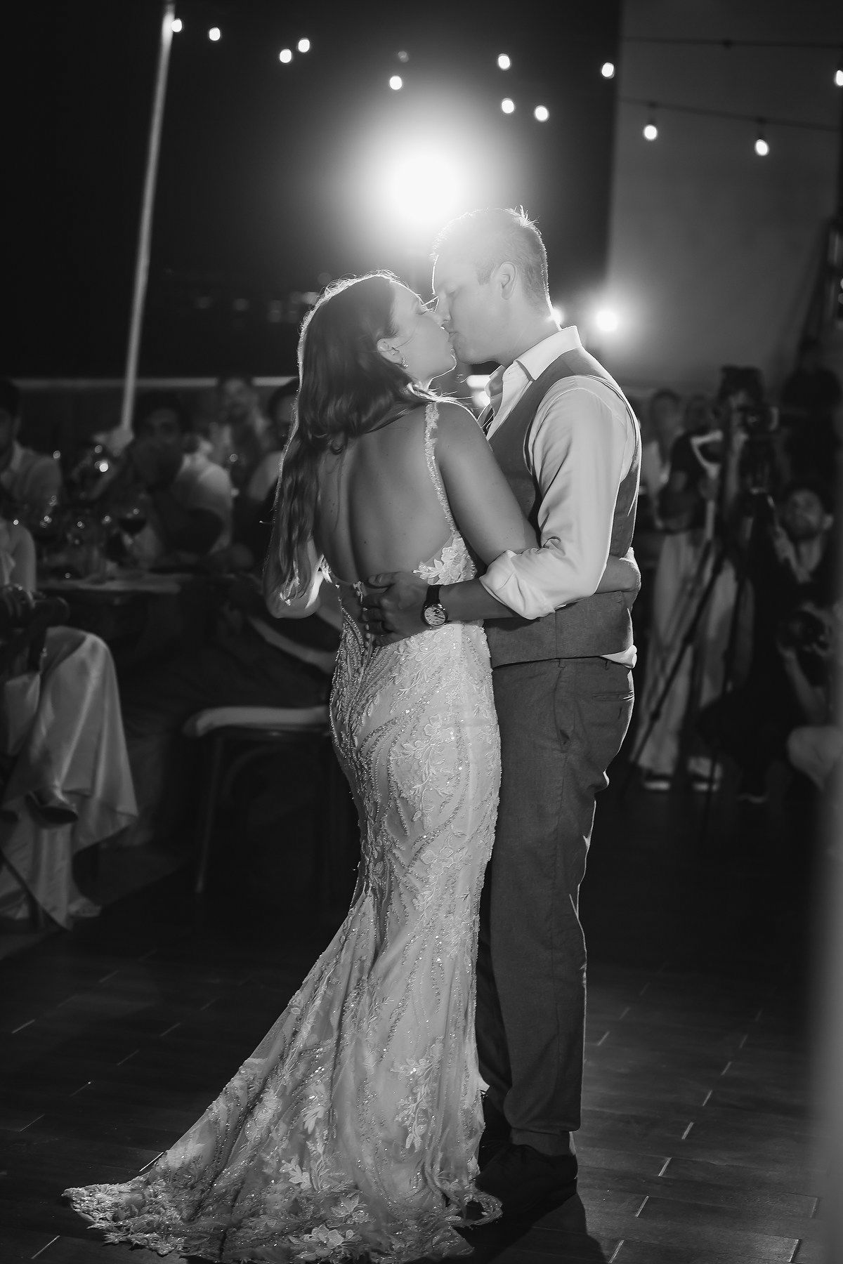 Black and white photo of a bride and groom sharing their first dance kiss at a romantic evening wedding reception.