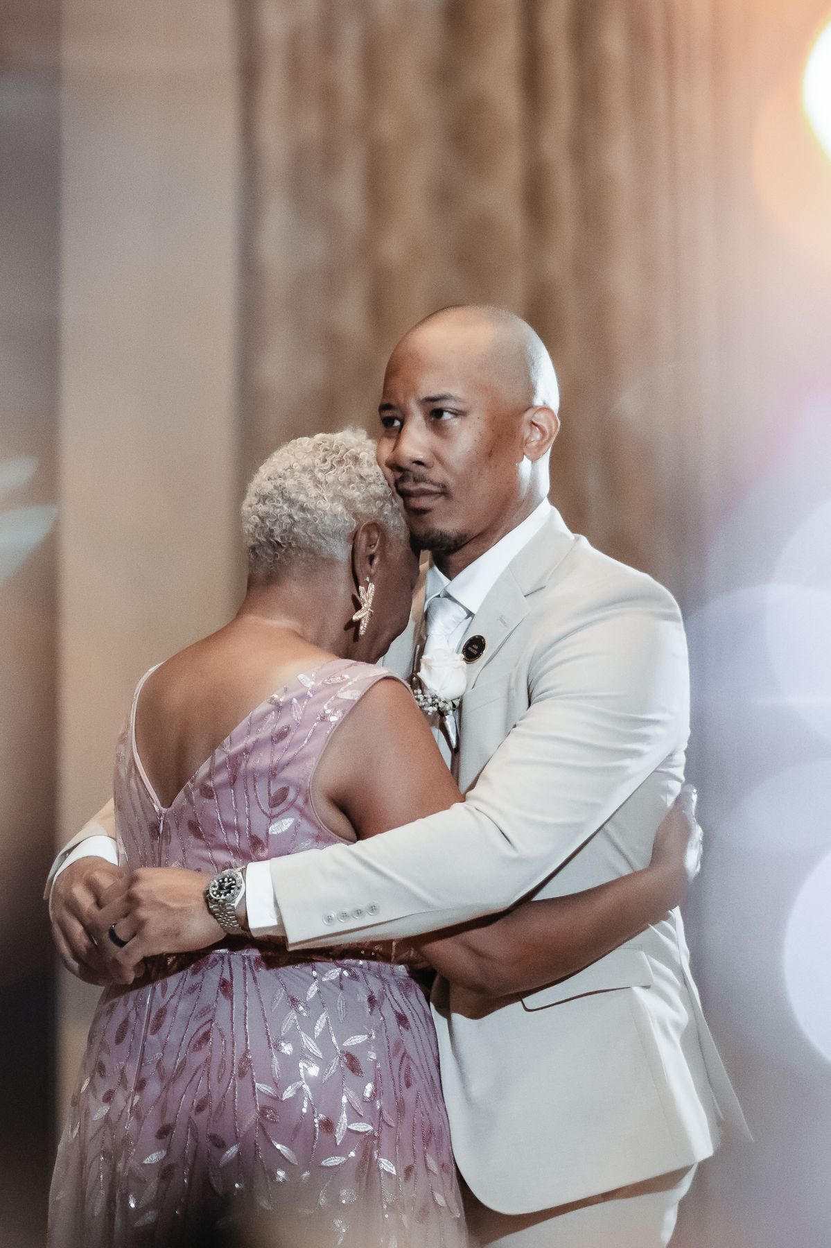 A groom in a white suit shares a tender dance with an older woman in a beaded lavender gown at a wedding reception.