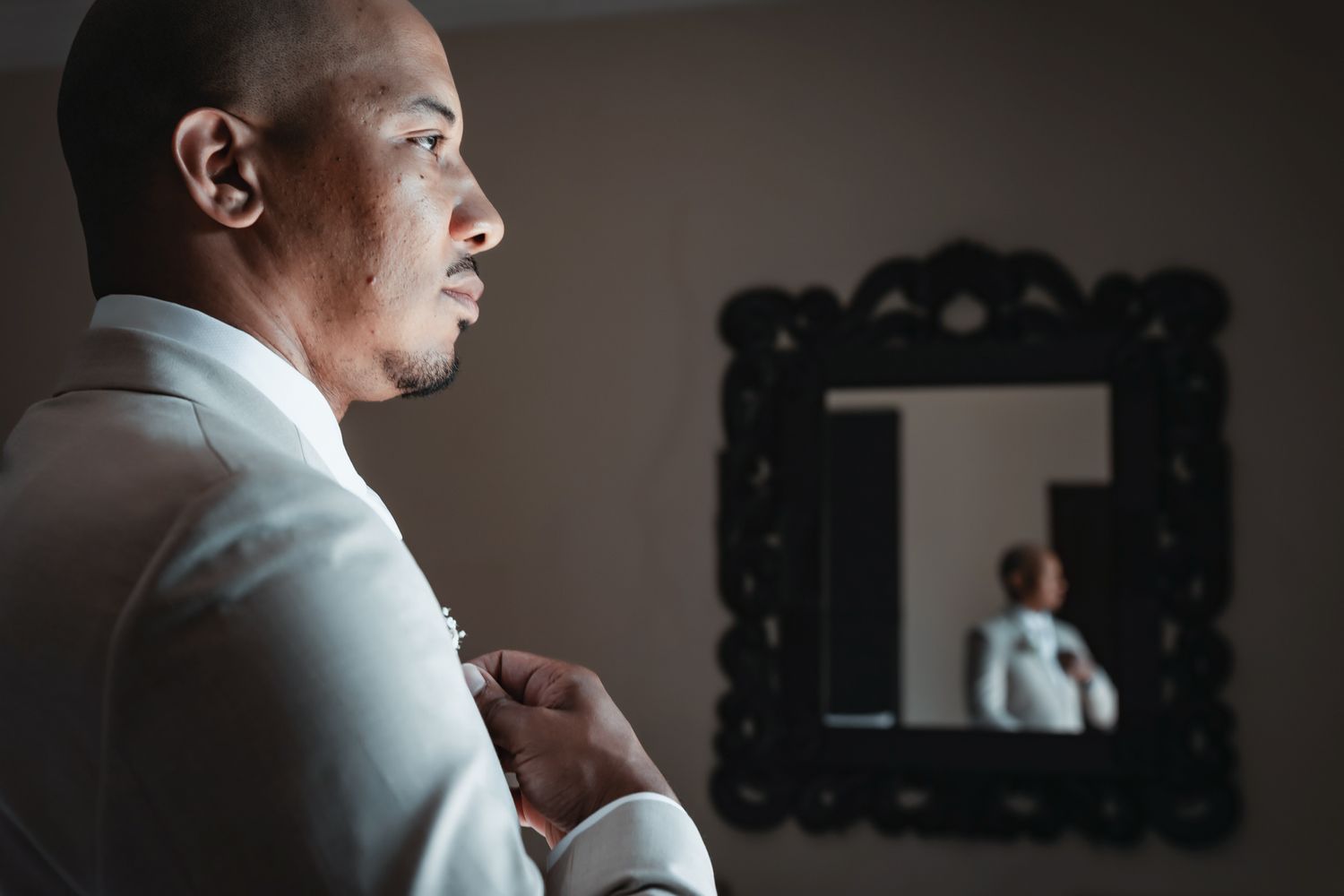 Groom in white suit adjusting boutonniere with ornate mirror reflection in background.