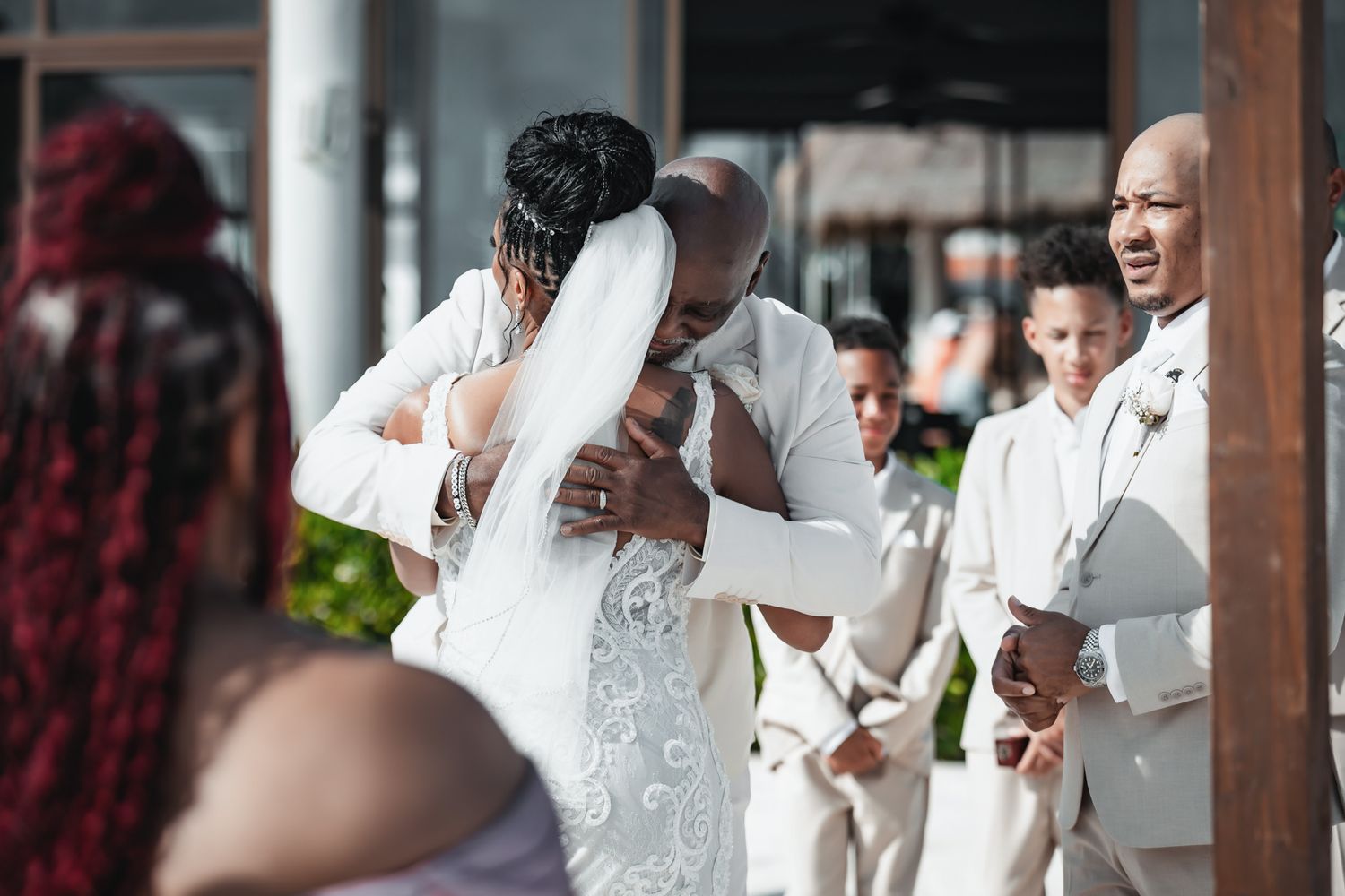 Groom embracing bride in white wedding dress and veil during outdoor wedding ceremony with groomsmen in background.