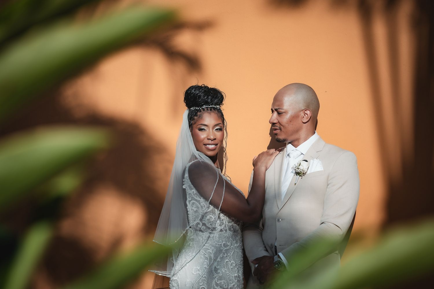 A bride in a beaded gown and veil adjusts her groom's boutonniere outdoors on their wedding day.