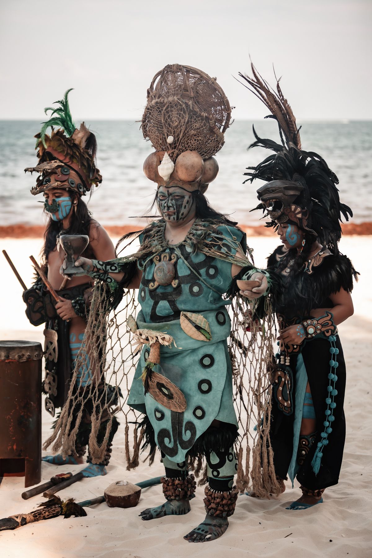 Three performers in elaborate Mayan tribal costumes with feathered headdresses on a sandy beach.