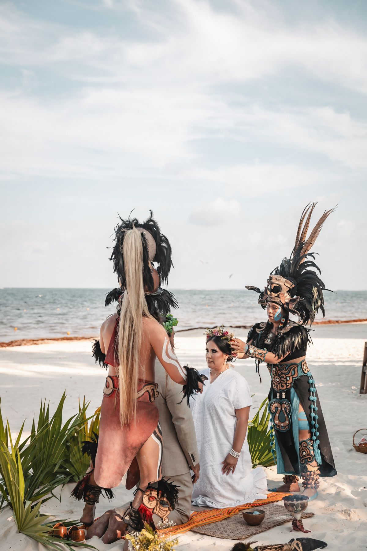Bride kneels during traditional Mayan beach ceremony surrounded by performers in black feathered headdresses by the ocean.