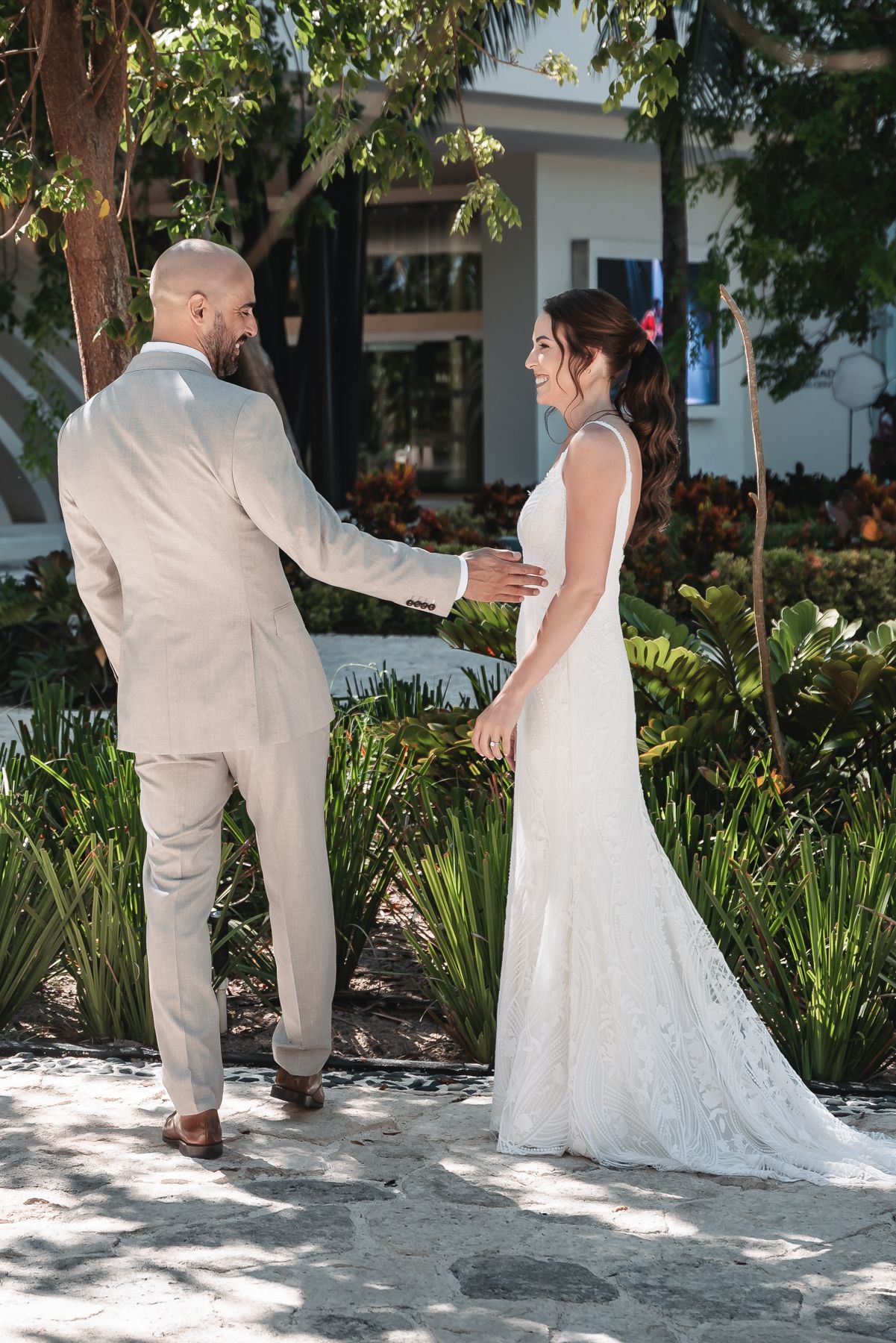 A bride in a white lace gown and groom in a tan suit share a romantic first look moment in a tropical garden.