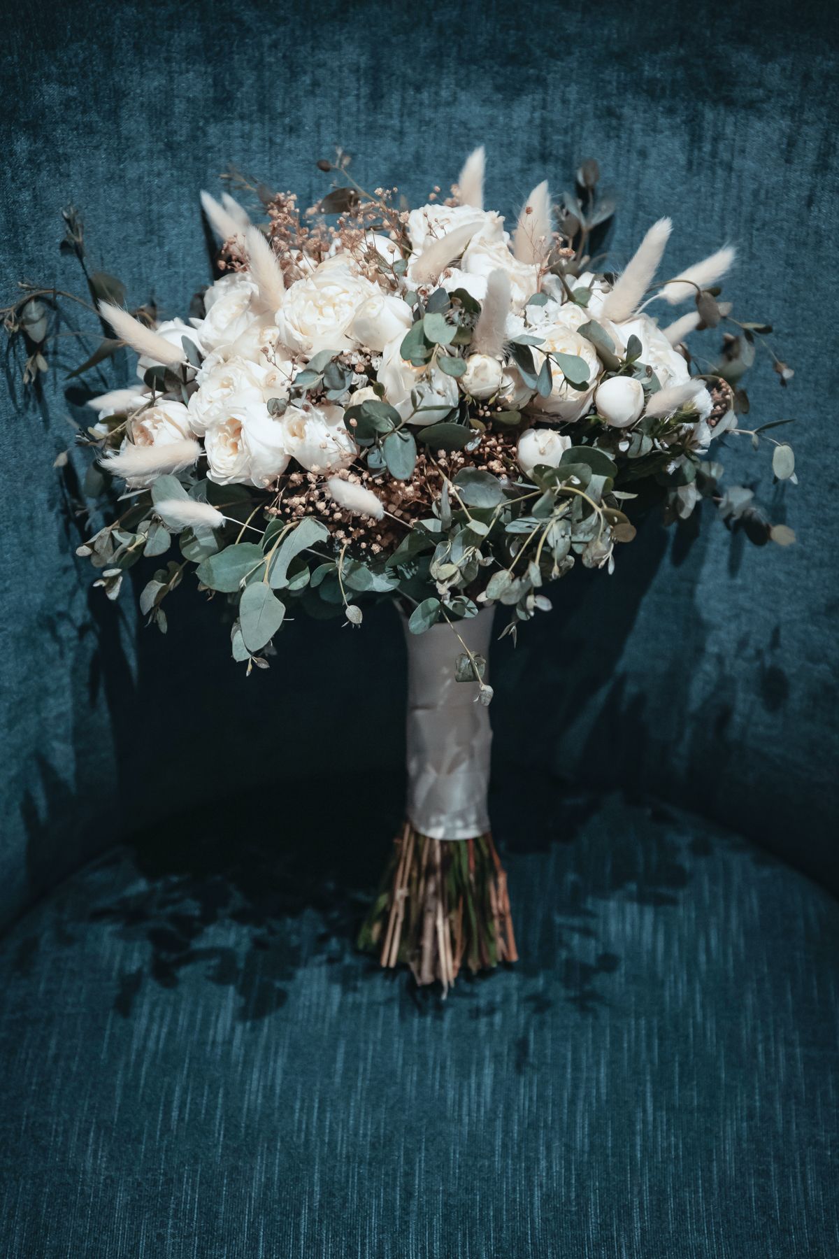 Elegant bridal bouquet with white roses, eucalyptus, and dried flowers wrapped in satin ribbon on blue velvet chair.