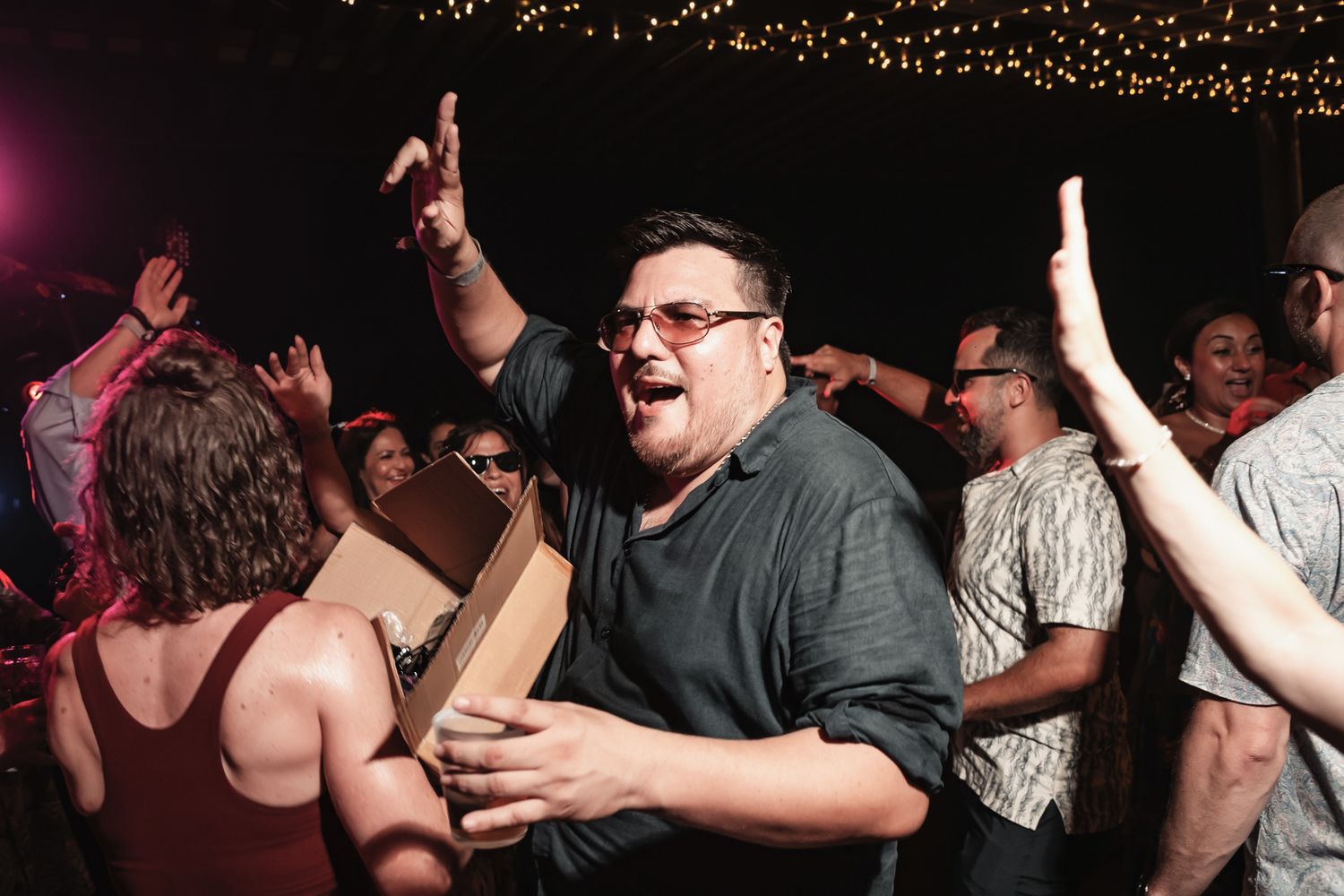 Excited crowd dancing at a lively nighttime outdoor party event with string lights in the background.