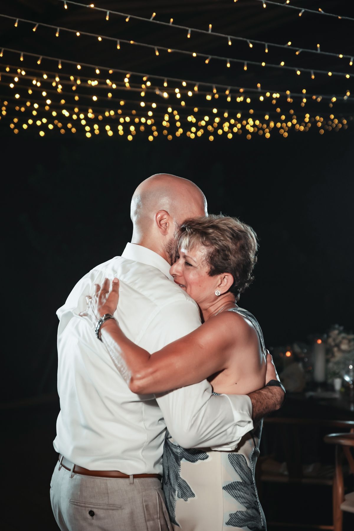 Couple sharing an emotional dance embrace under warm string lights at an outdoor evening wedding reception.