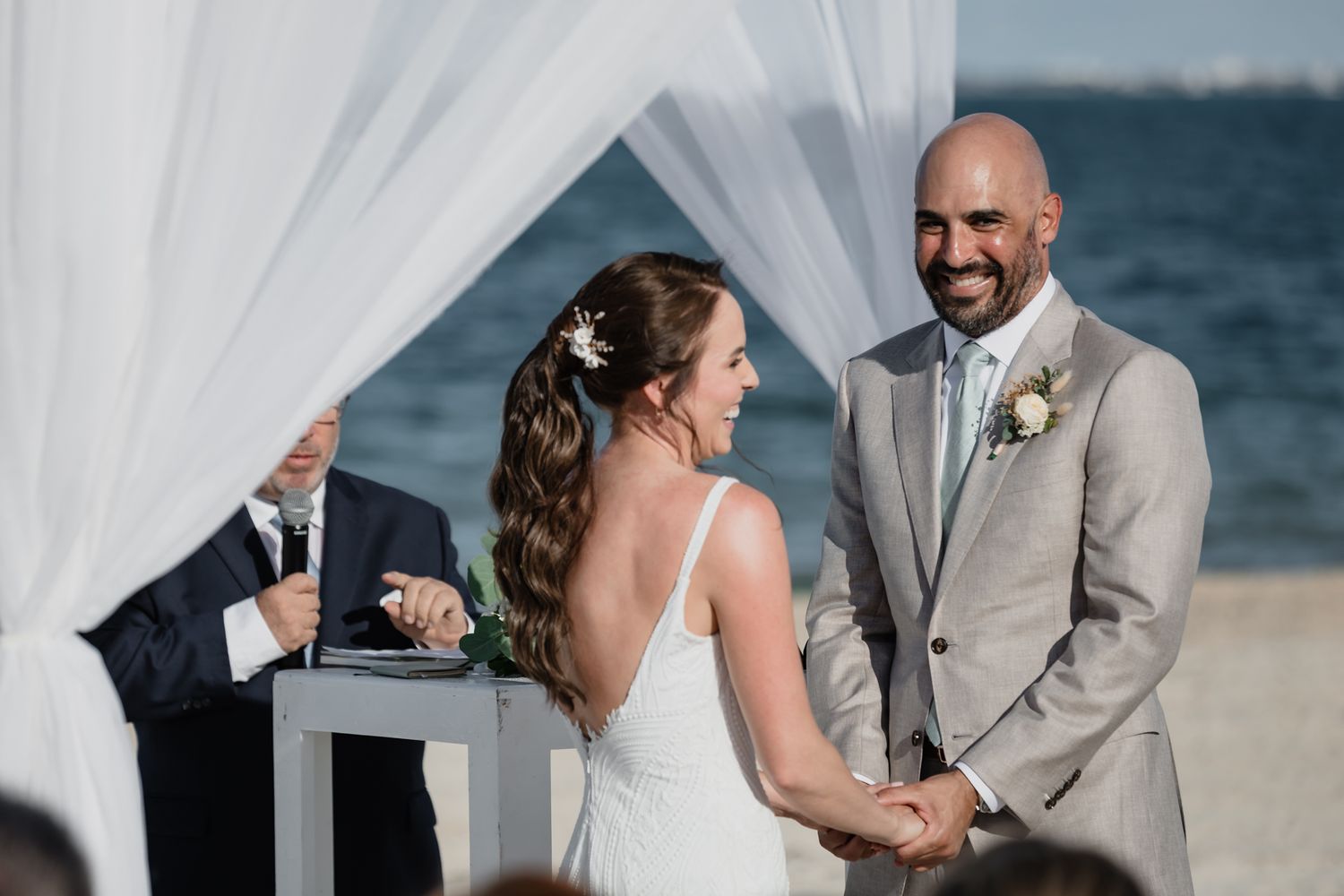 Bride and groom exchanging vows at a beach wedding ceremony under white draping with ocean backdrop.
