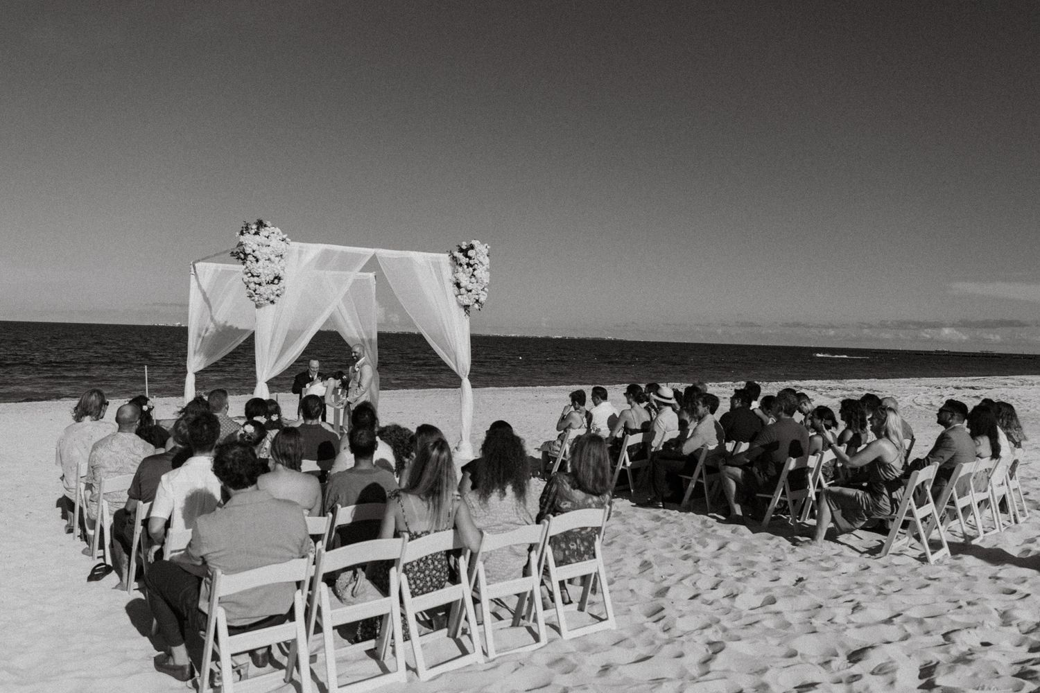 Black and white beach wedding ceremony with guests seated on white chairs facing a floral arch by the ocean.