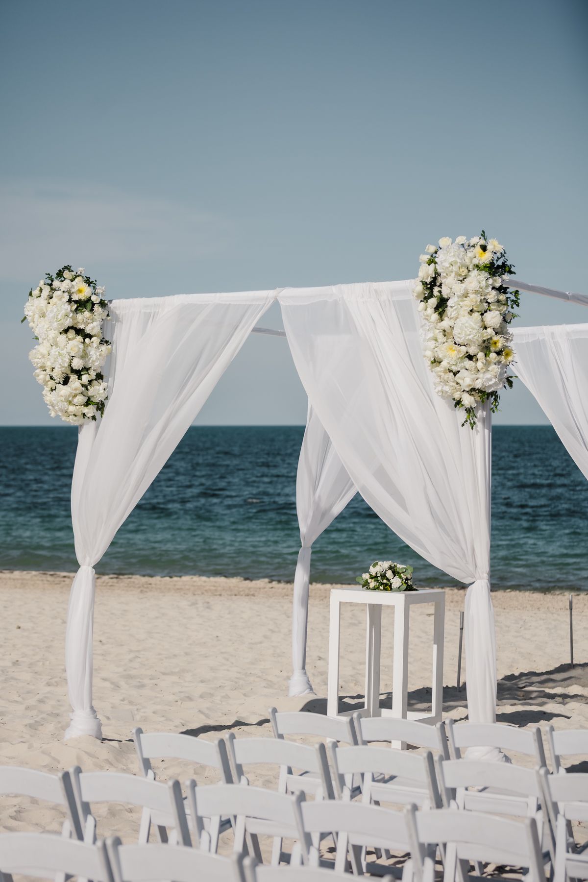 White draped beach wedding arch adorned with white floral arrangements, white chairs, and ocean backdrop.