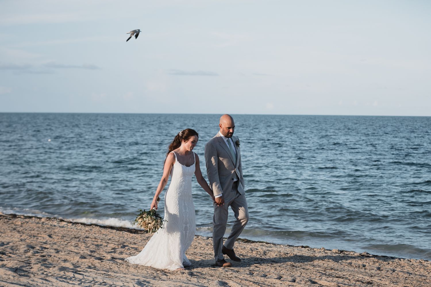 Bride and groom walking hand in hand on a sandy beach by the ocean on their wedding day.