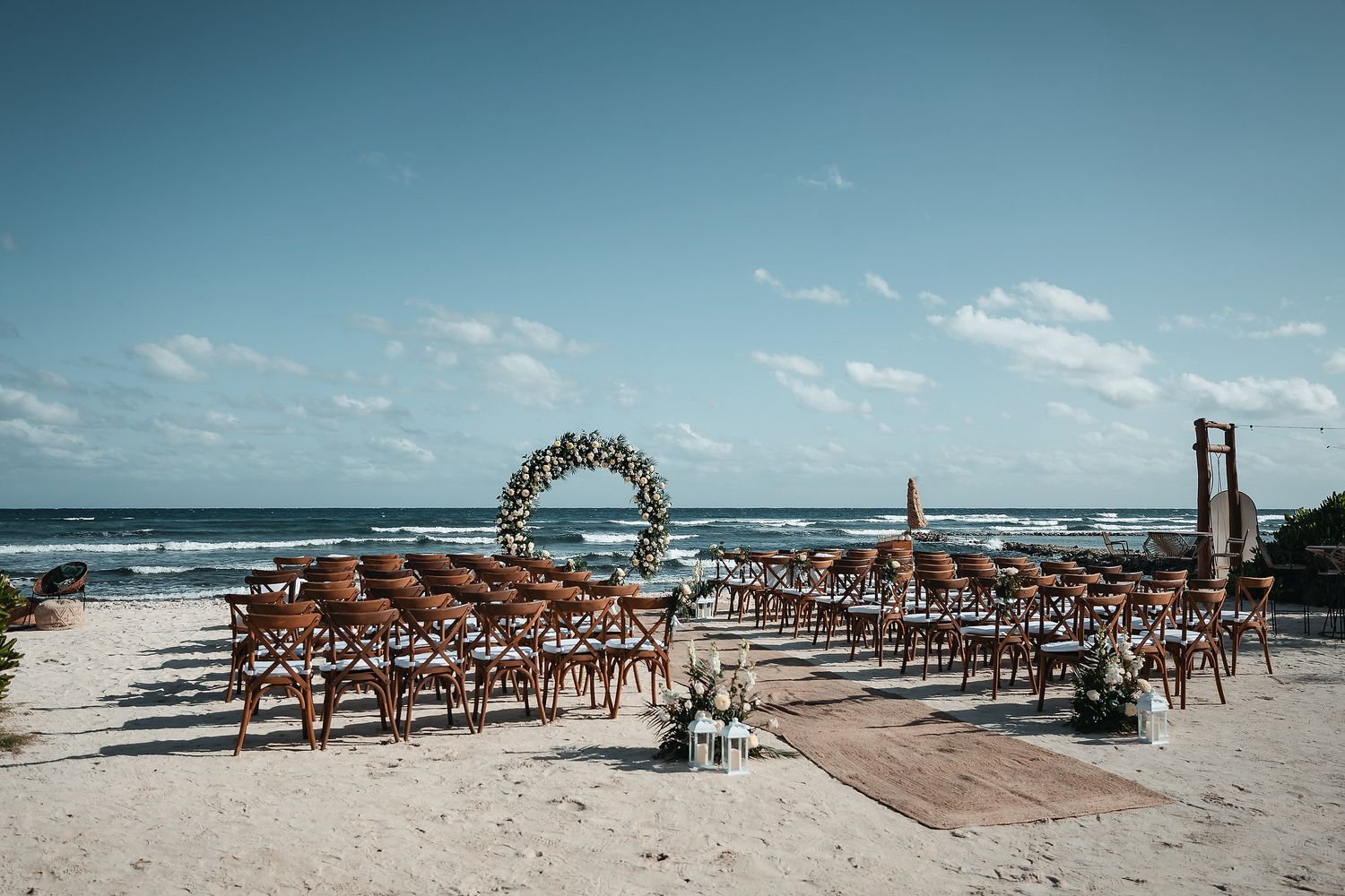 Elegant beach wedding ceremony setup with wooden chairs, floral arch, and ocean backdrop on a sunny day.