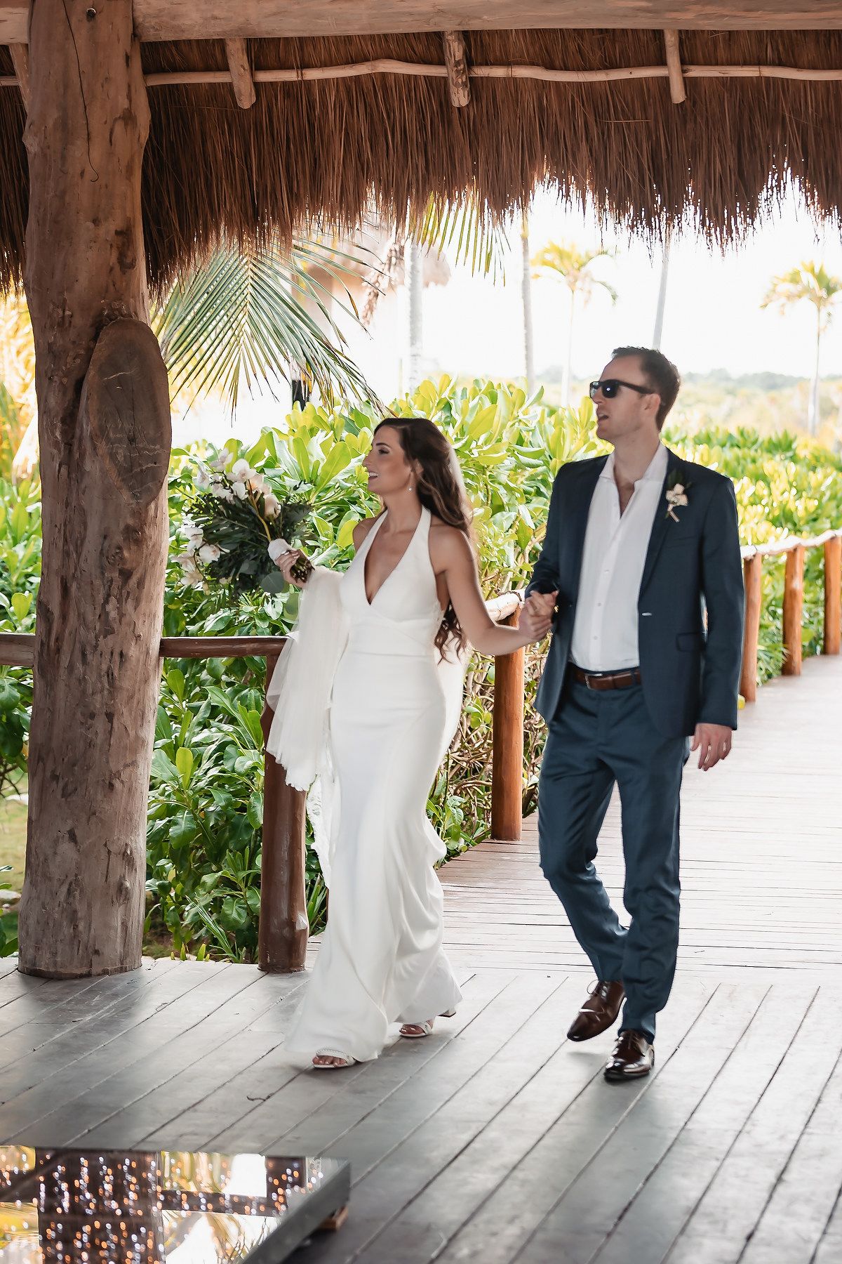 Bride and groom walking hand-in-hand under a thatched palapa roof surrounded by tropical greenery.