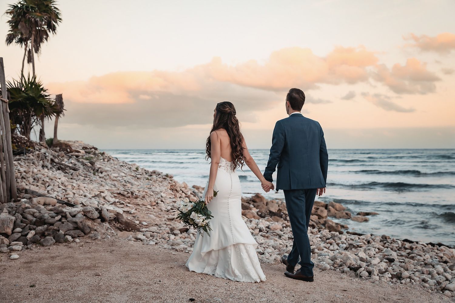 Newlywed couple holding hands walking along rocky beach at sunset, bride in white gown, groom in navy suit.
